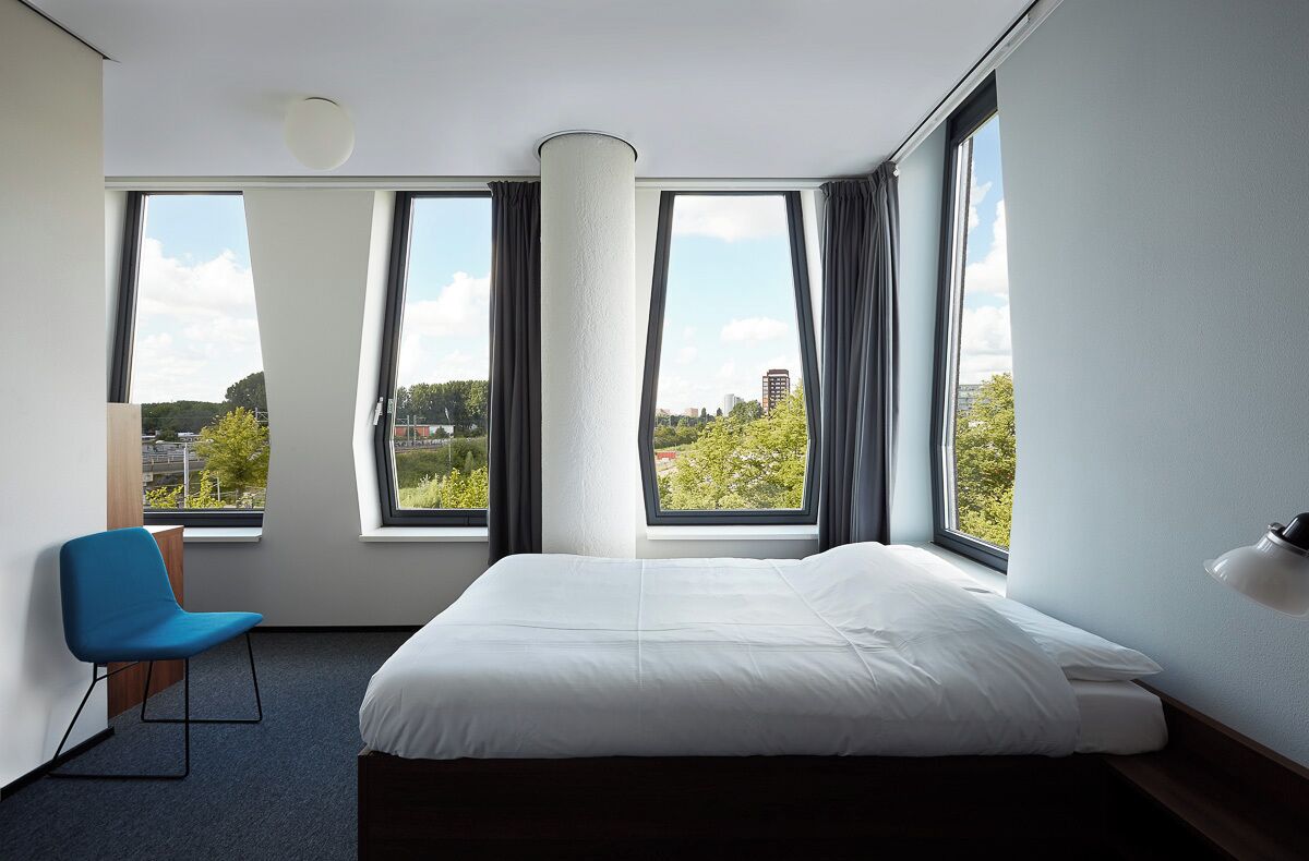 Habitación de hotel con una cama, una silla, ventanas grandes y vistas de la ciudad.