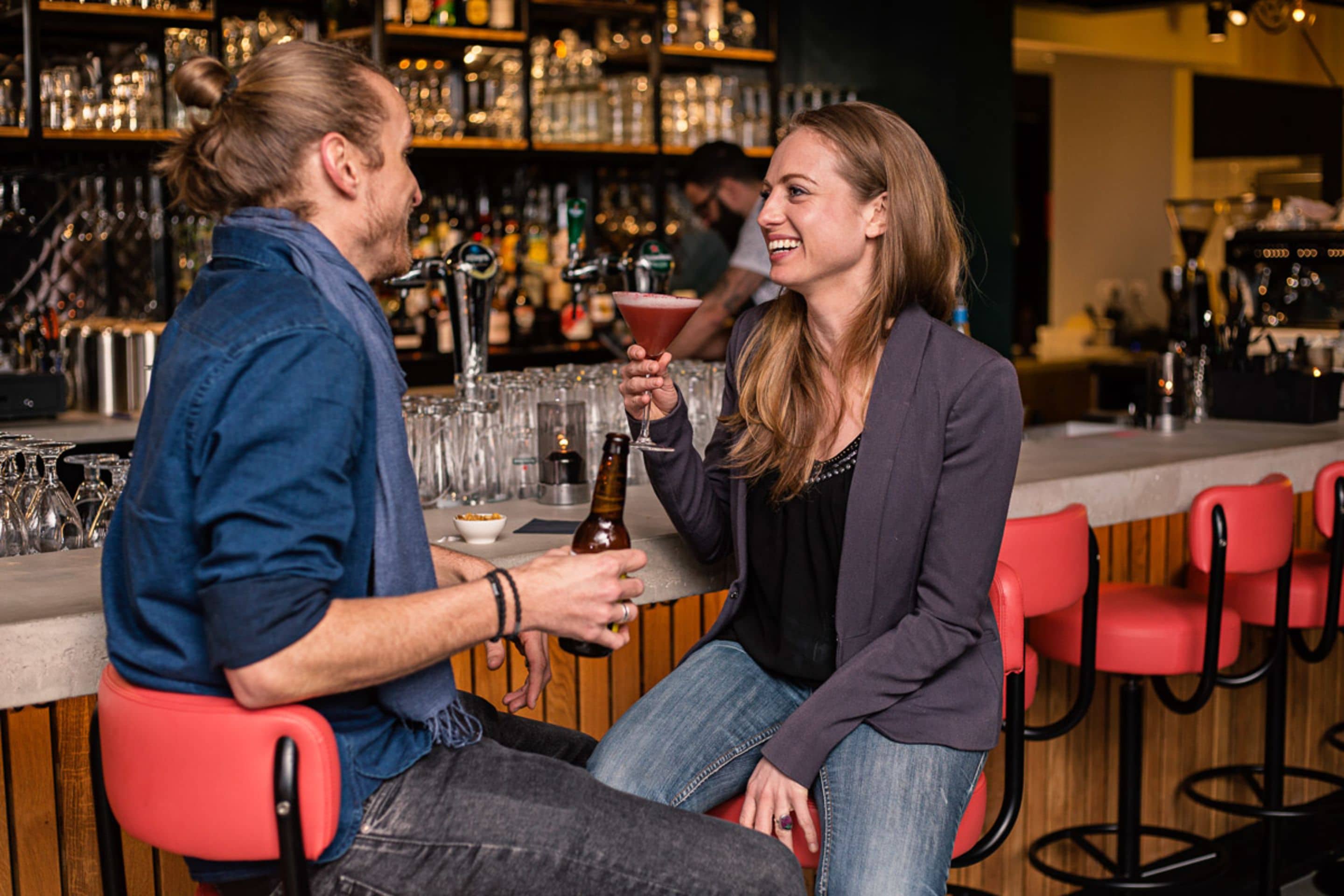 Twee personen zitten aan een bar, met verschillende flessen en glazen.