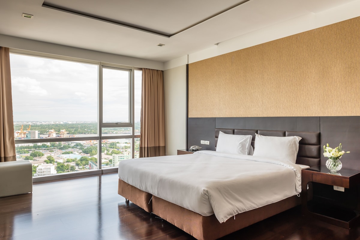 A modern hotel room with a large bed, a nightstand, and a view of a cityscape through large windows.