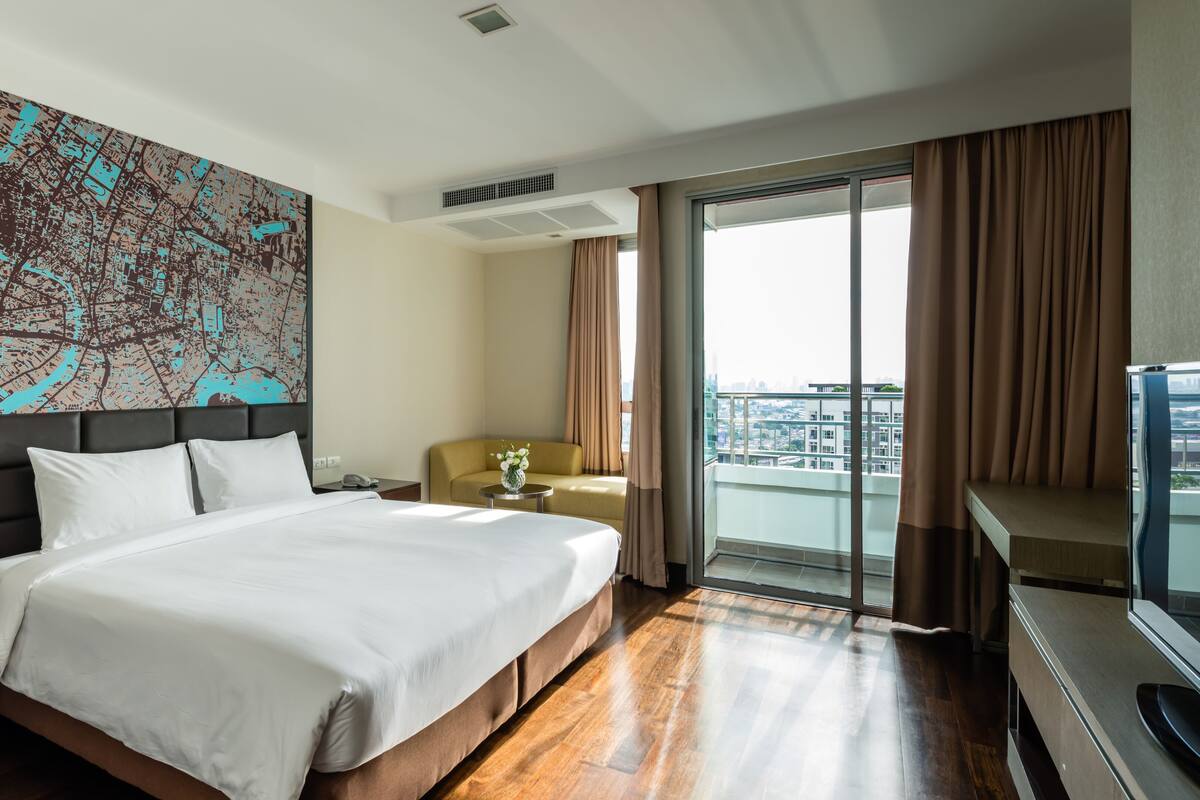 A hotel room with a large bed, a cityscape artwork on the wall, a sofa, a small table with a vase, and a balcony with curtains.