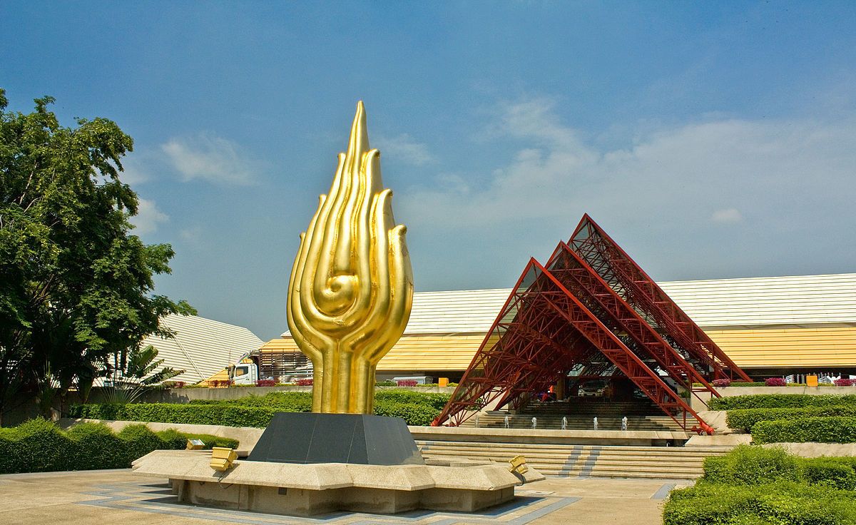 Een gouden vlamsculptuur in een aangelegd gebied, met een modern gebouw en een rode trap op de achtergrond.