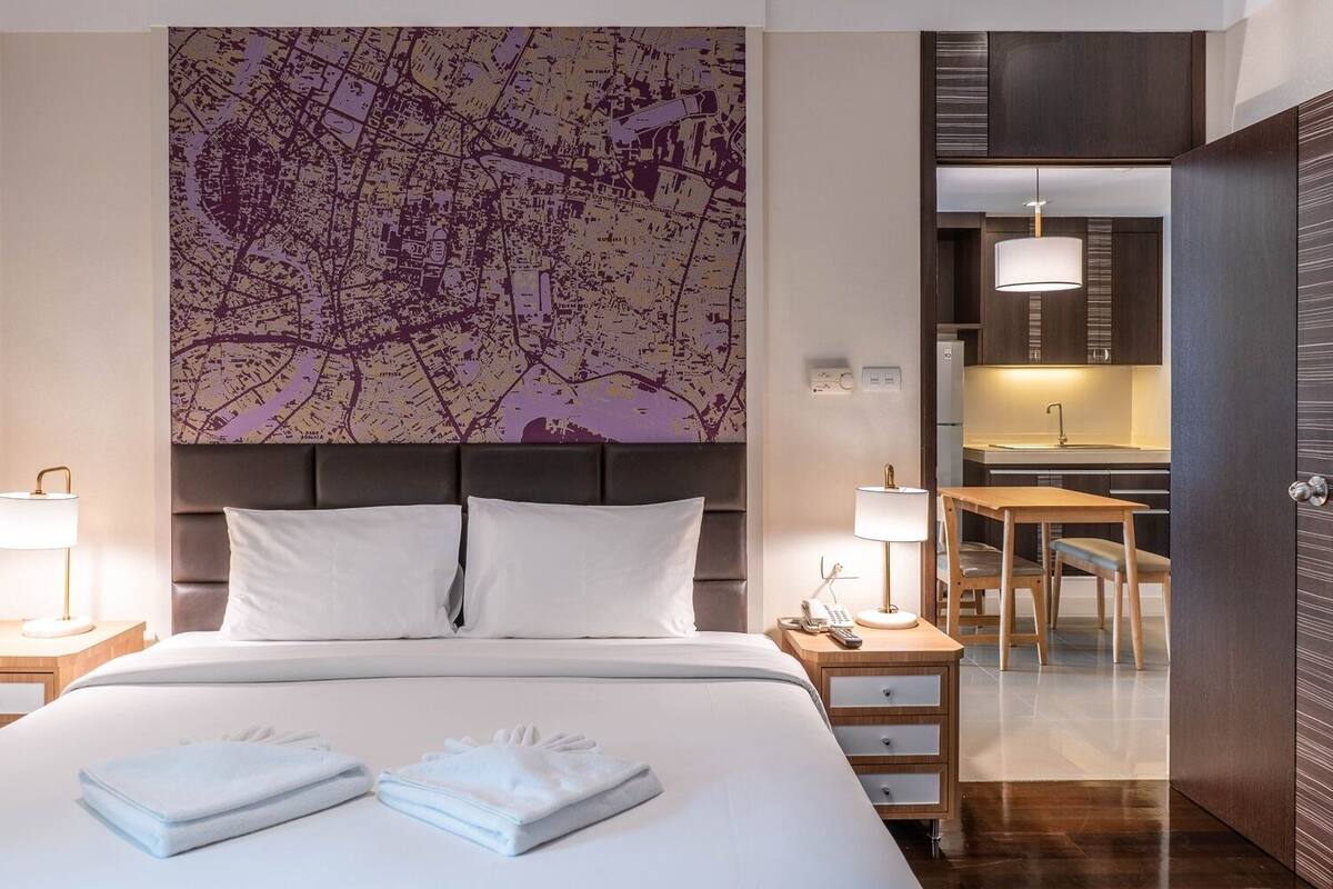 A hotel room with a large bed, a map artwork on the wall, a desk with a chair, and a kitchenette.