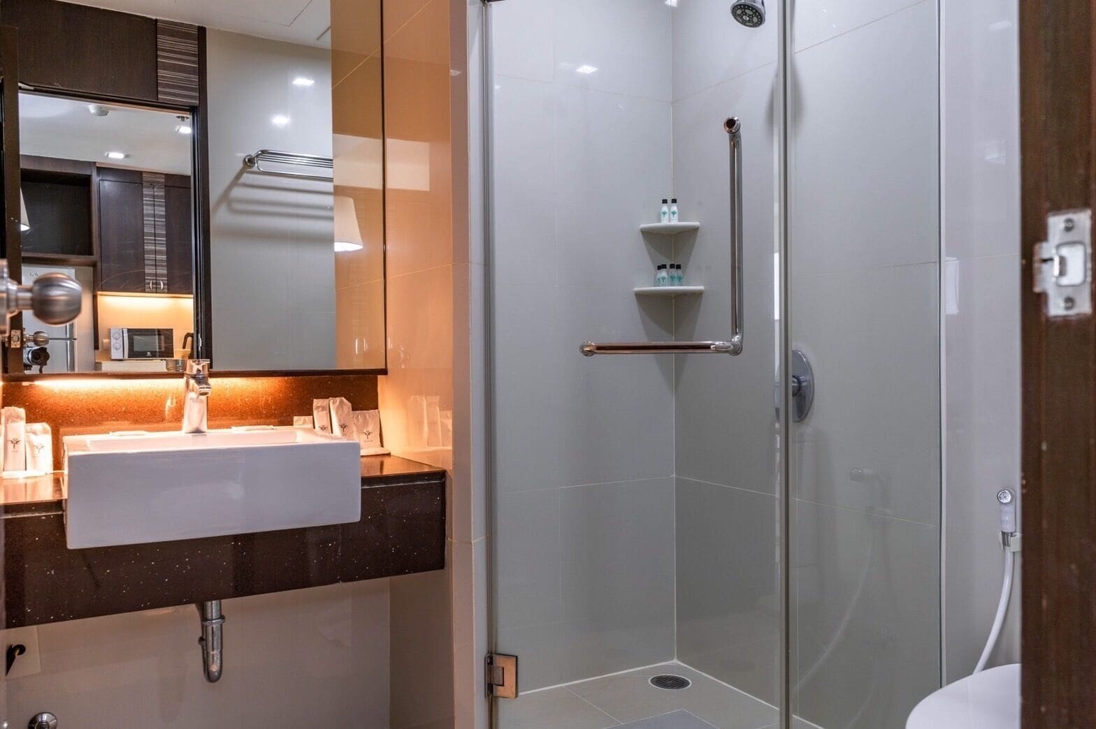 Un baño moderno con ducha envuelta en vidrio, un lavamanos blanco con un espejo encima y un estante pequeño con artículos de aseo.