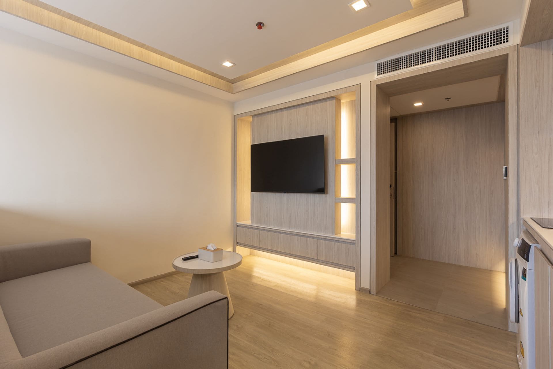 A modern hotel room with a sofa, a small round table, a flat-screen TV mounted on a wooden panel, and a wardrobe with a built-in heater.