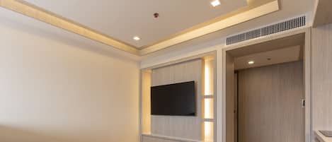 A modern hotel room with a sofa, a small round table, a flat-screen TV mounted on a wooden panel, and a wardrobe with a built-in heater.
