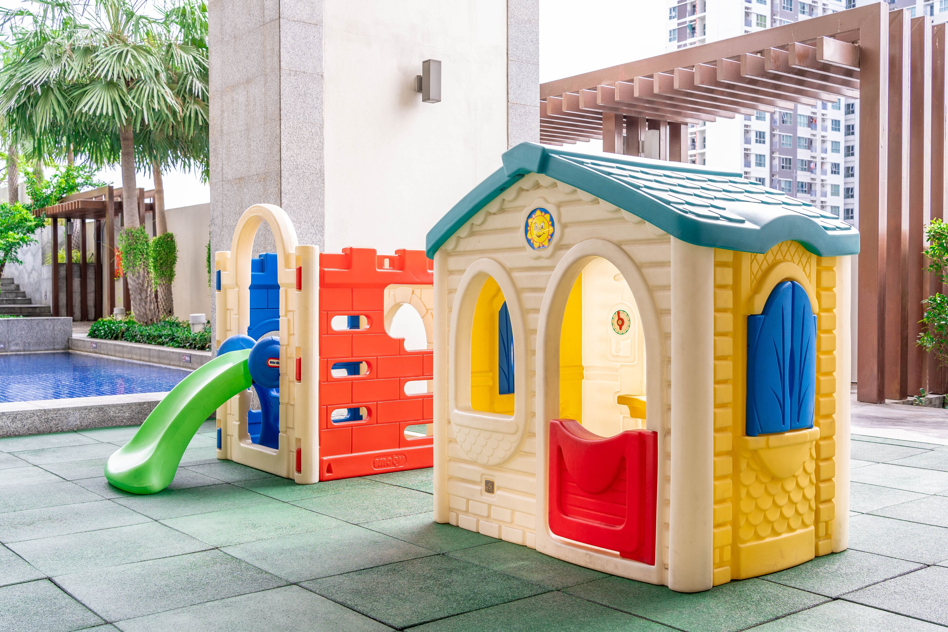 A children's play area with a slide and two playhouses, one red and one yellow, near a pool.