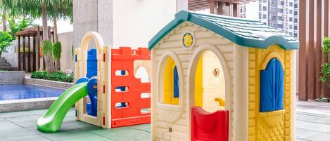 Zona infantil con un tobogán y dos casitas de juegos, una roja y otra amarilla, cerca de una piscina.