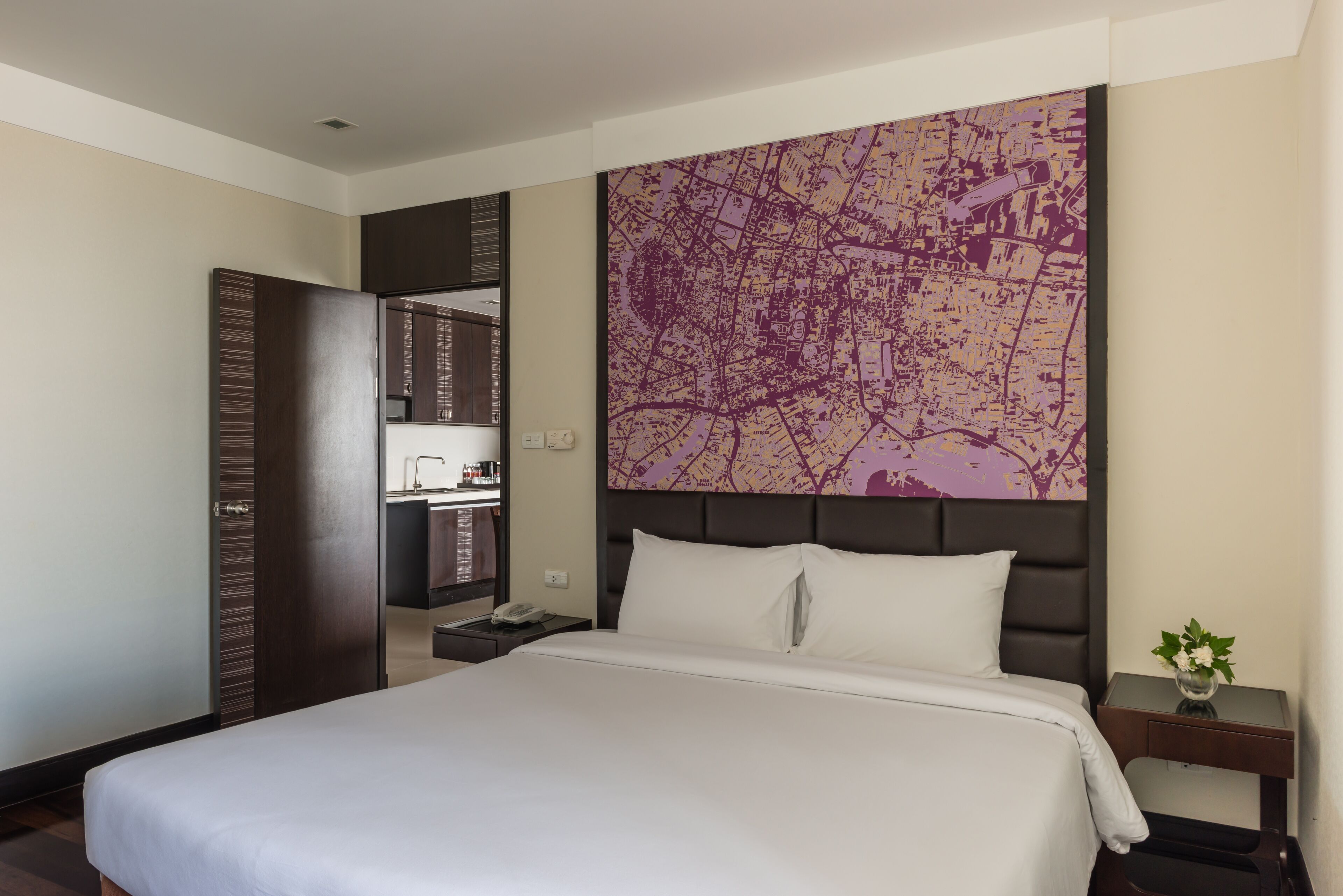 A hotel room with a large bed, a map on the wall, a small table with a vase, and a bathroom visible through an open door.