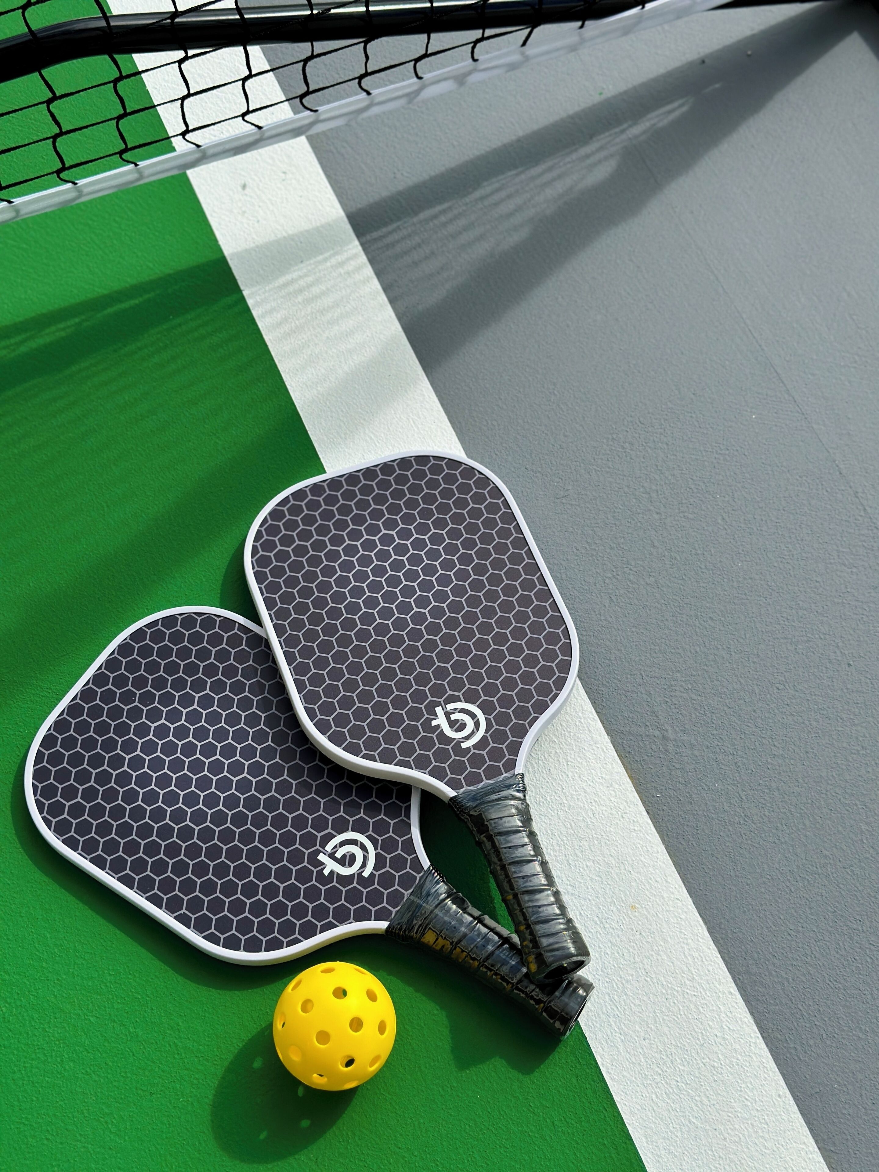 Dos paletas de tenis de mesa y una pelota sobre una cancha verde y gris con líneas blancas.