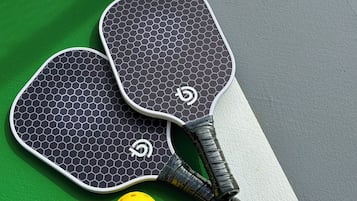 Dos paletas de tenis de mesa y una pelota sobre una cancha verde y gris con líneas blancas.