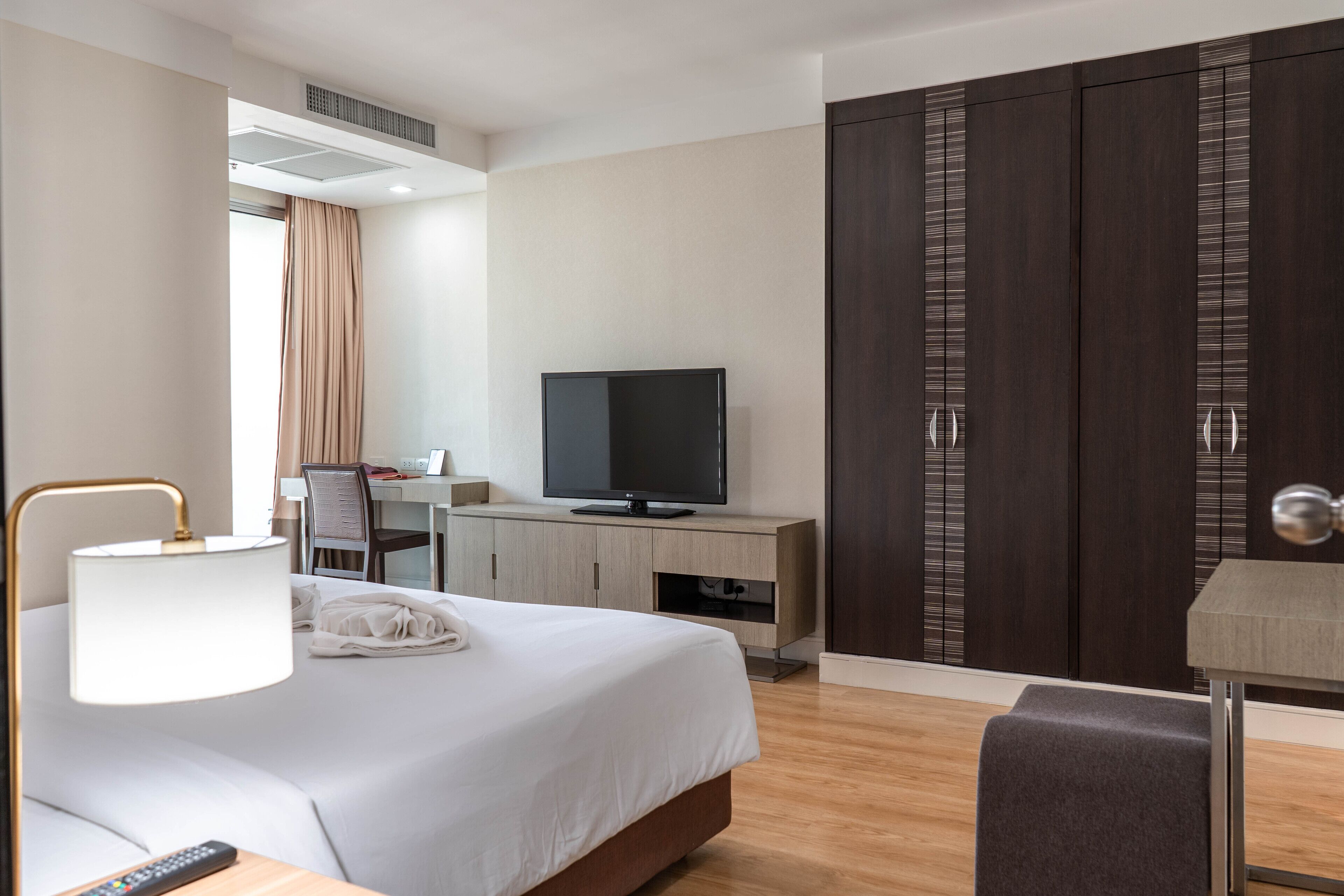A modern hotel room with a bed, bedside table, lamp, TV, wardrobe, desk, and chair.