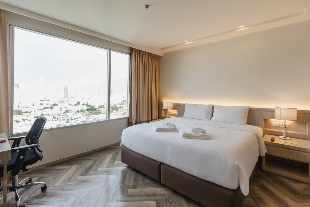 A hotel room with a large bed, a desk, and a chair. There is a view of a cityscape through the window.