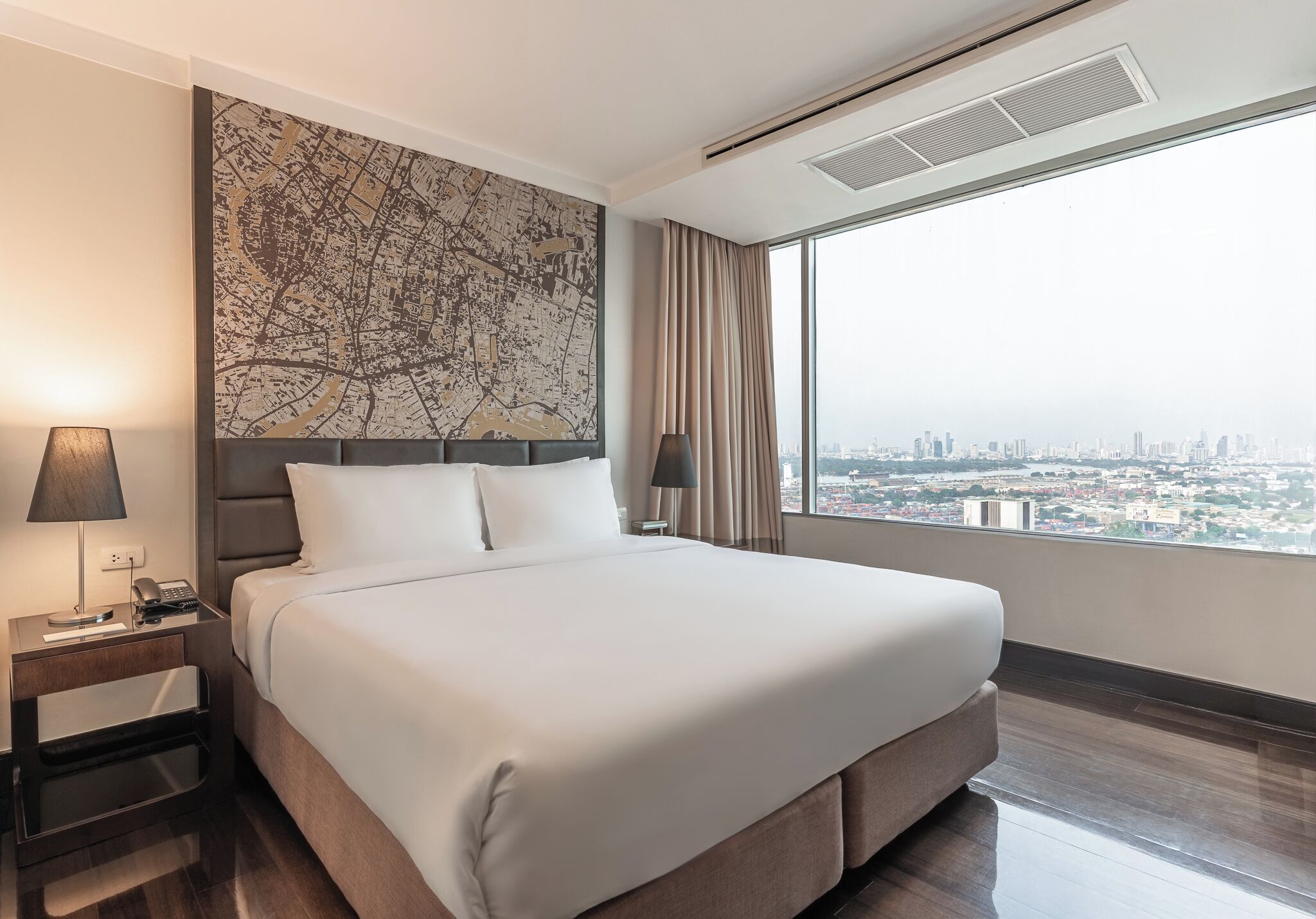A hotel room with a large bed, a nightstand with a lamp, a cityscape view through a large window, and a map artwork on the wall.