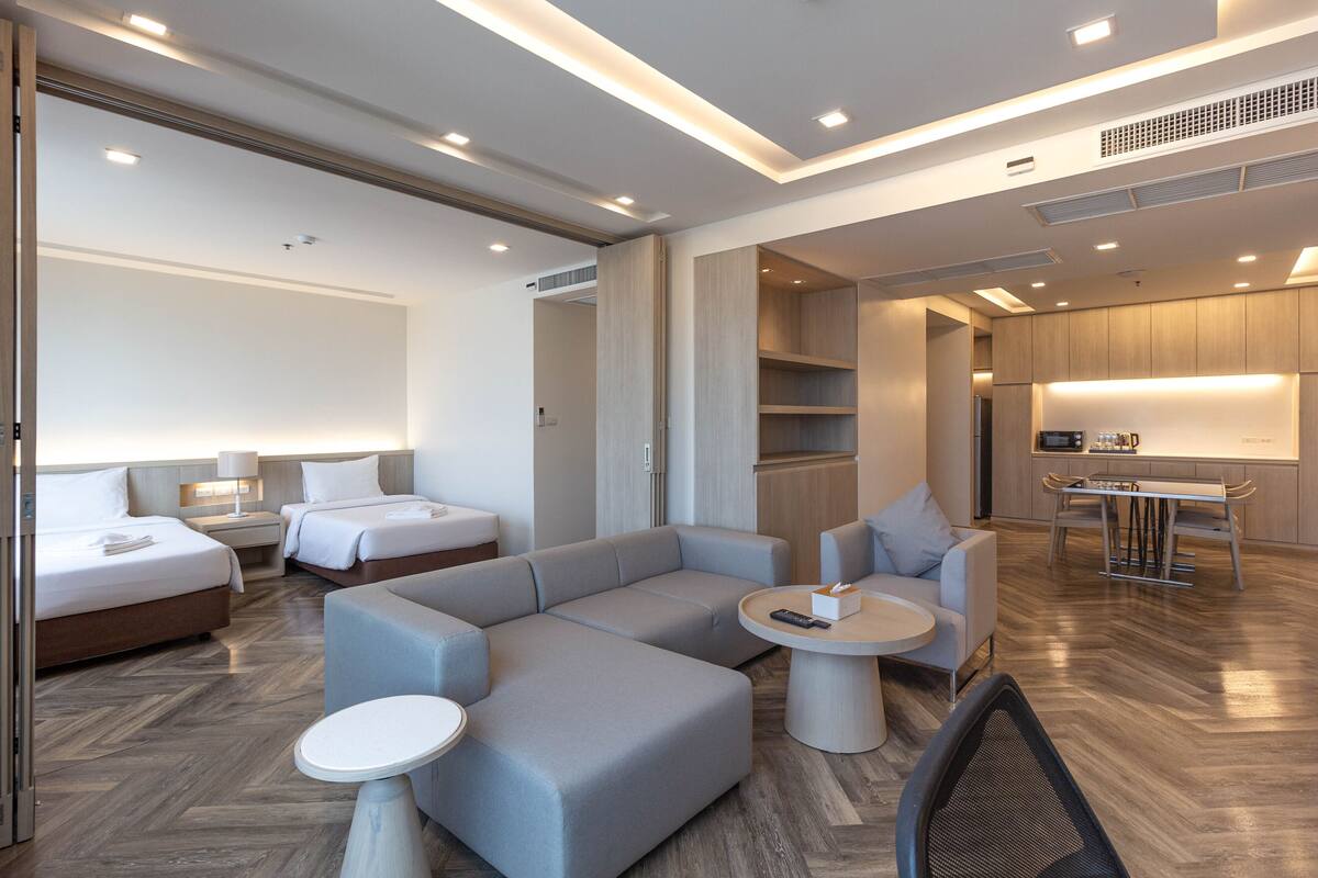 A modern hotel room with a large L-shaped sofa, two single beds, a dining area, and a kitchenette.
