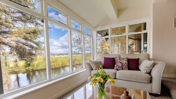 A room with a large window overlooking a river, a sofa with cushions, and a vase with flowers on a table. - Killarney View House B&B (Killarney)