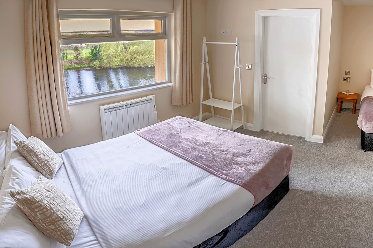 A bedroom with a large bed, a window with a view of a river, a white door, and a small table with a chair.