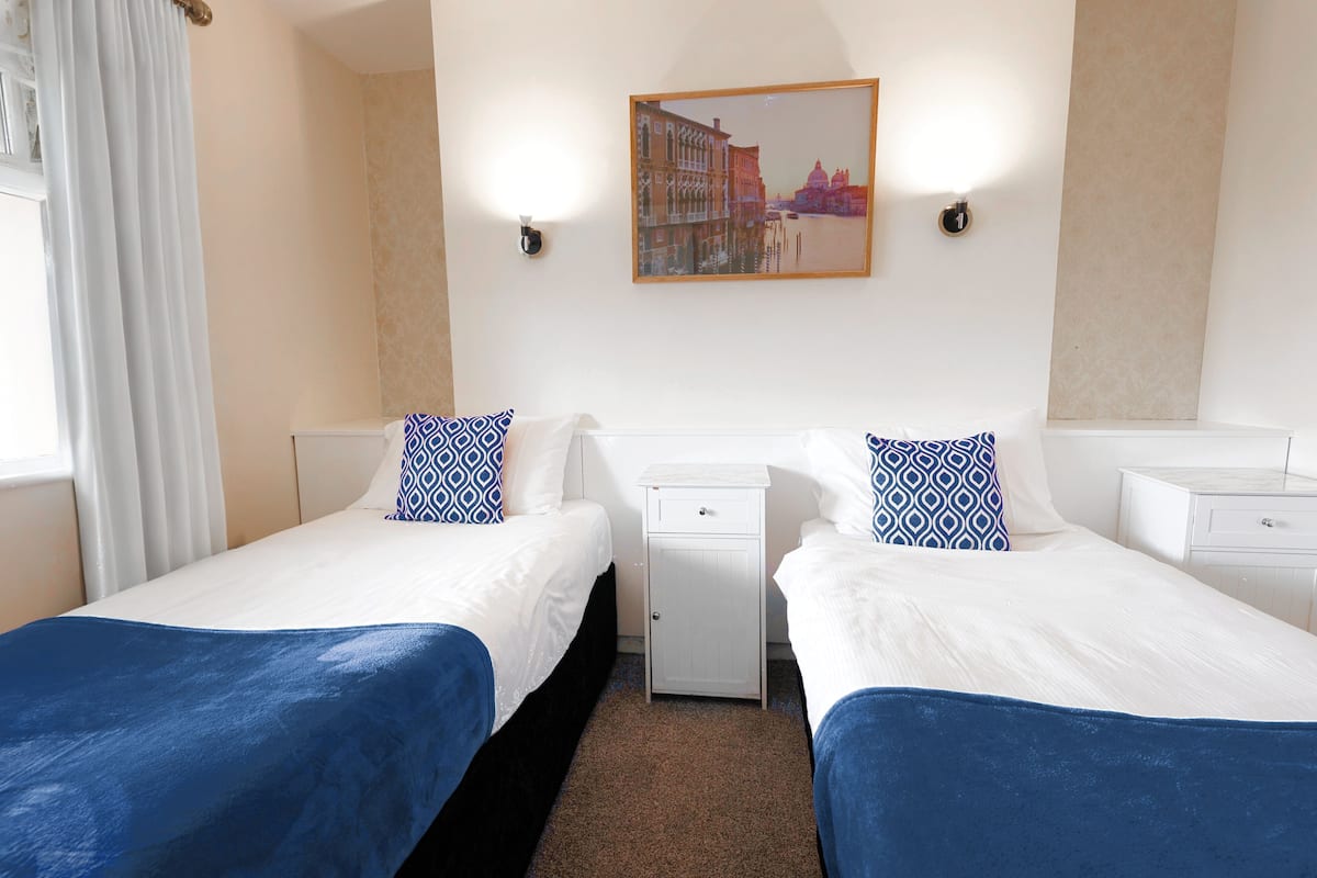A hotel room with two beds, a small bedside table, a nightstand, a framed picture on the wall, and a window with curtains.