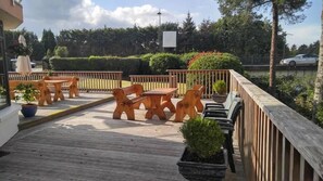 Une terrasse en bois, agrémentée de mobilier d’extérieur, notamment des tables et des chaises, entourée de verdure et d’une clôture.