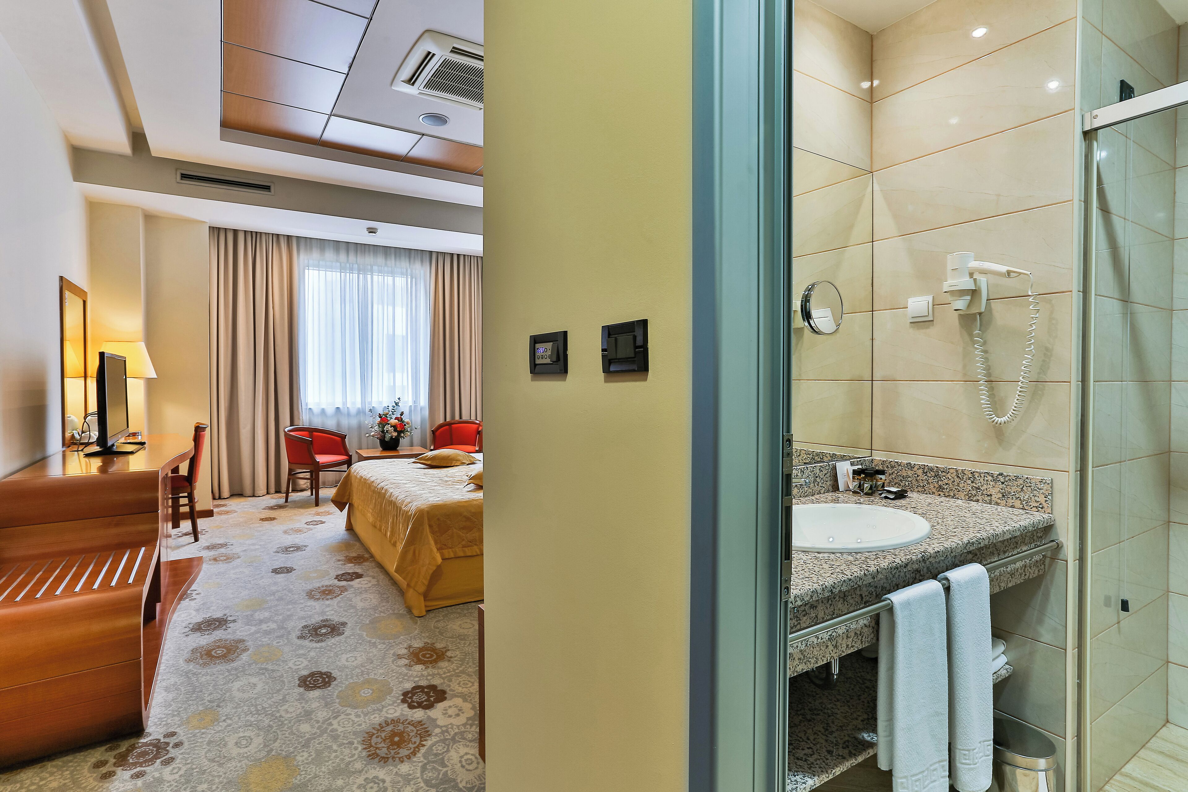 A hotel room with a bed, a desk, a chair, a lamp, and a bathroom with a sink and shower.