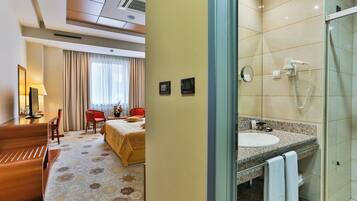 A hotel room with a bed, a desk, a chair, a lamp, and a bathroom with a sink and shower.