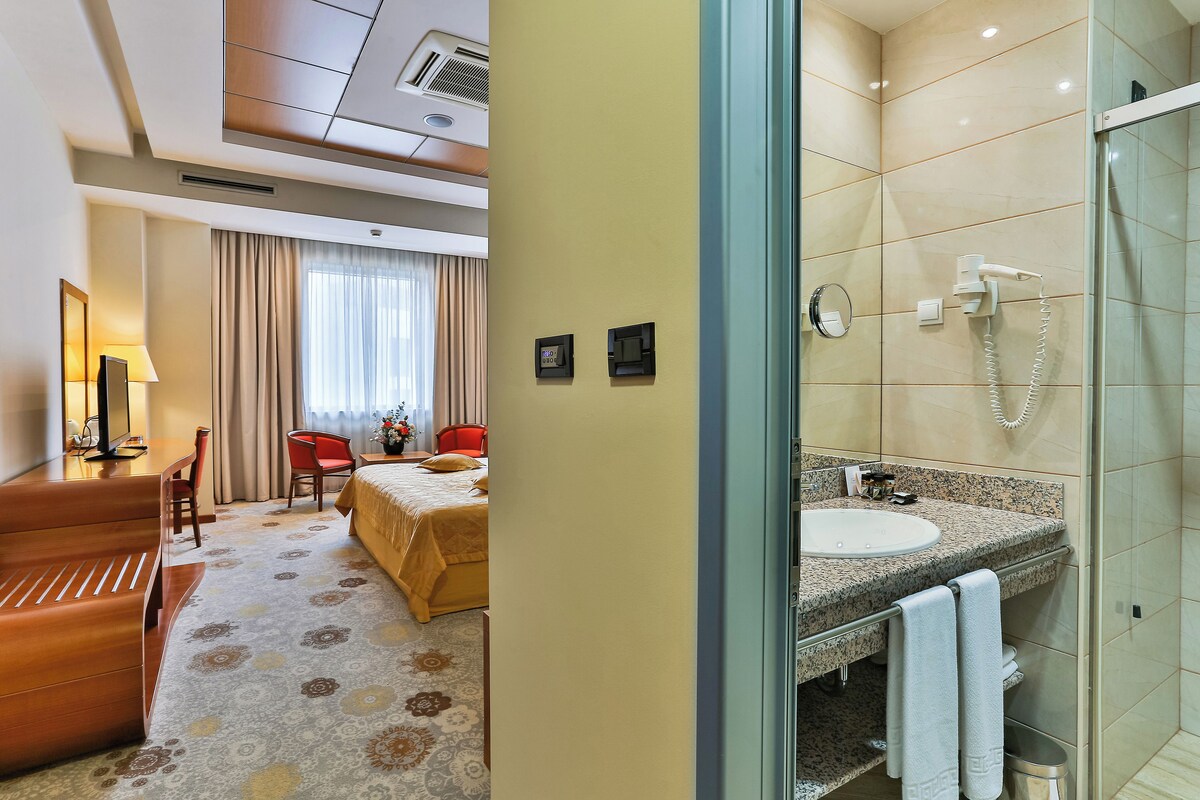 Habitación de hotel con cama, escritorio, silla, lámpara y baño con lavabo y ducha.