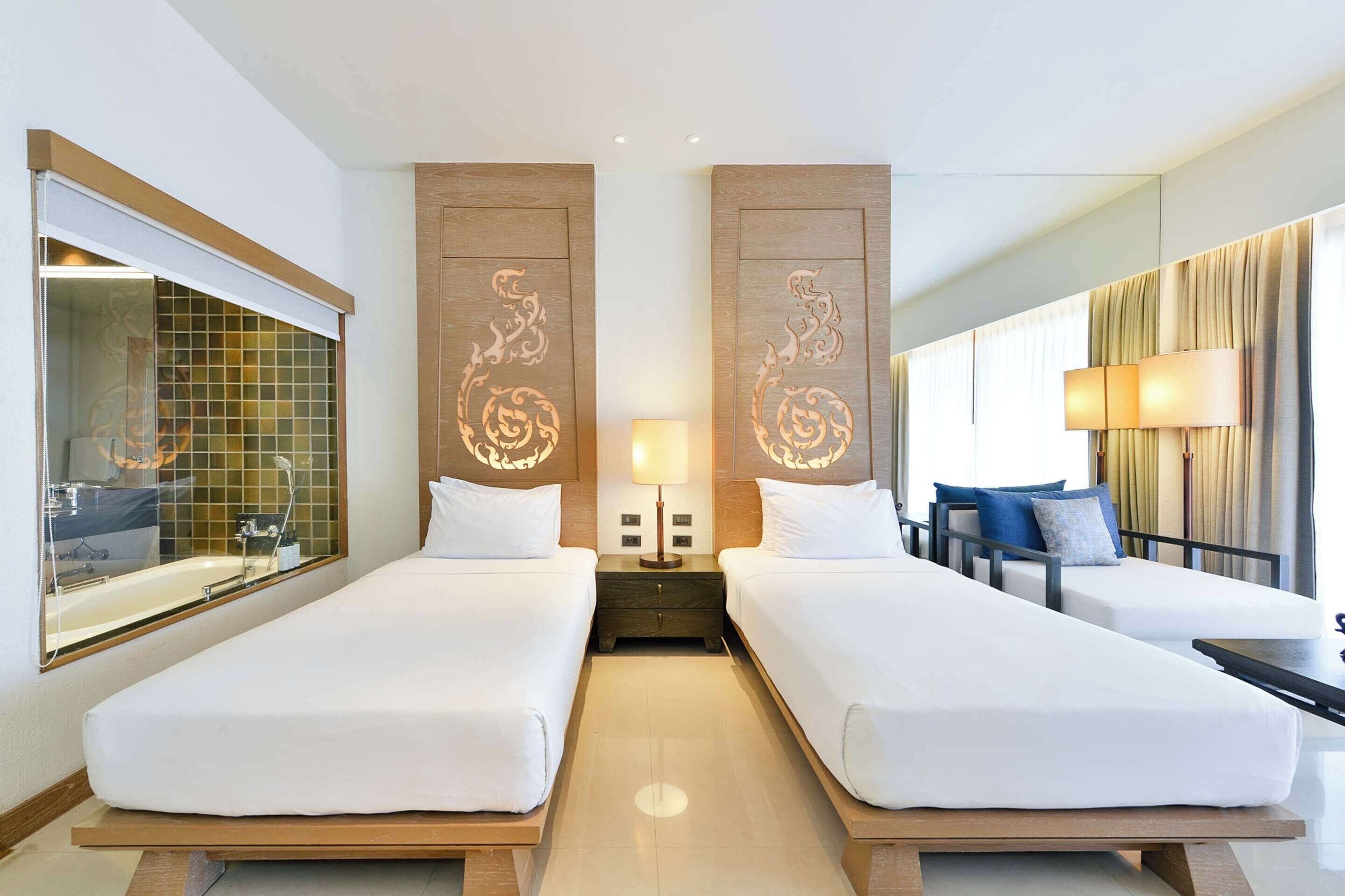 A hotel room with two beds, a nightstand with a lamp, a mirror, a window with curtains, and a view of the outside.
