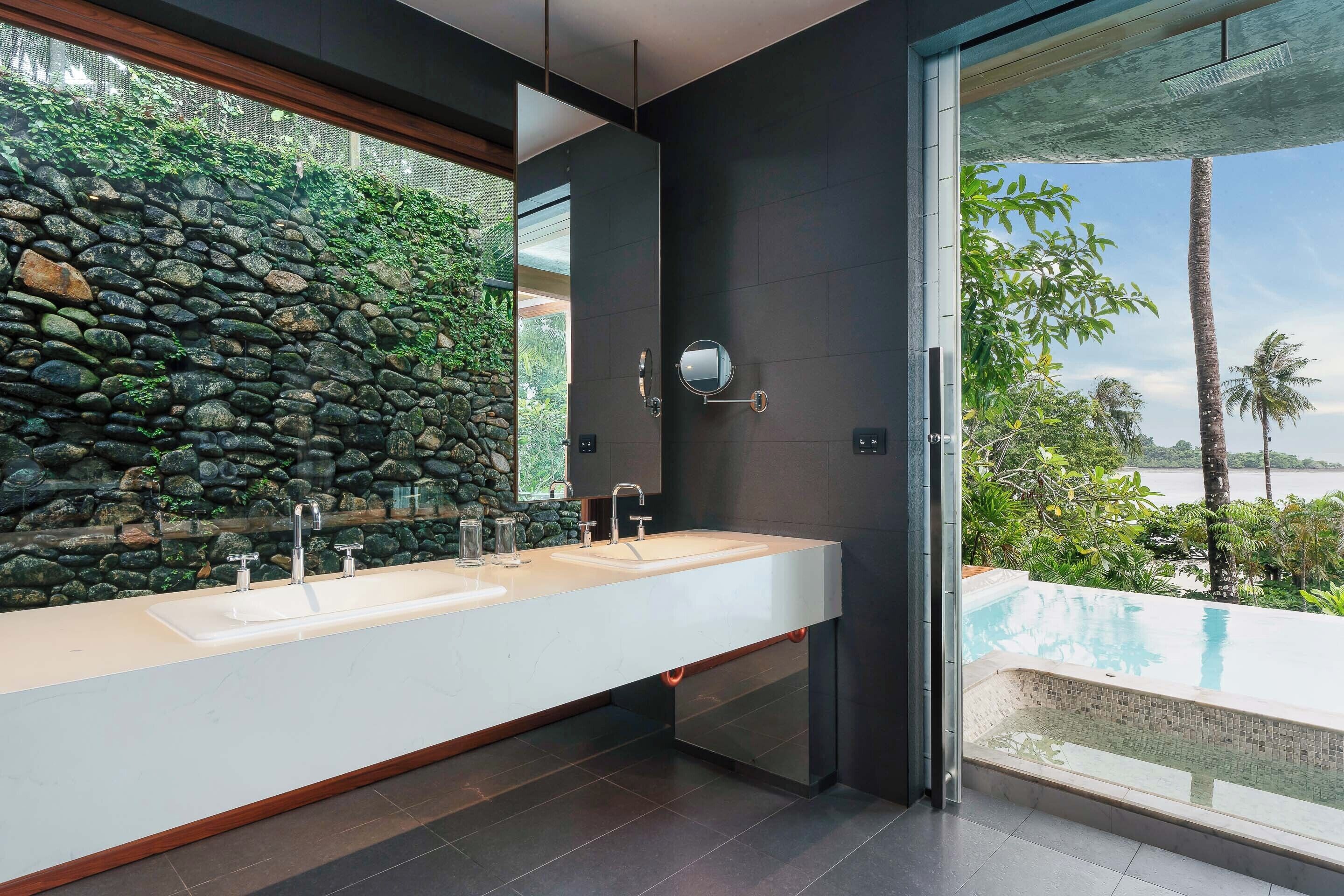 Une salle de bain moderne avec un mur en pierre, deux vasques et une vue sur une piscine.