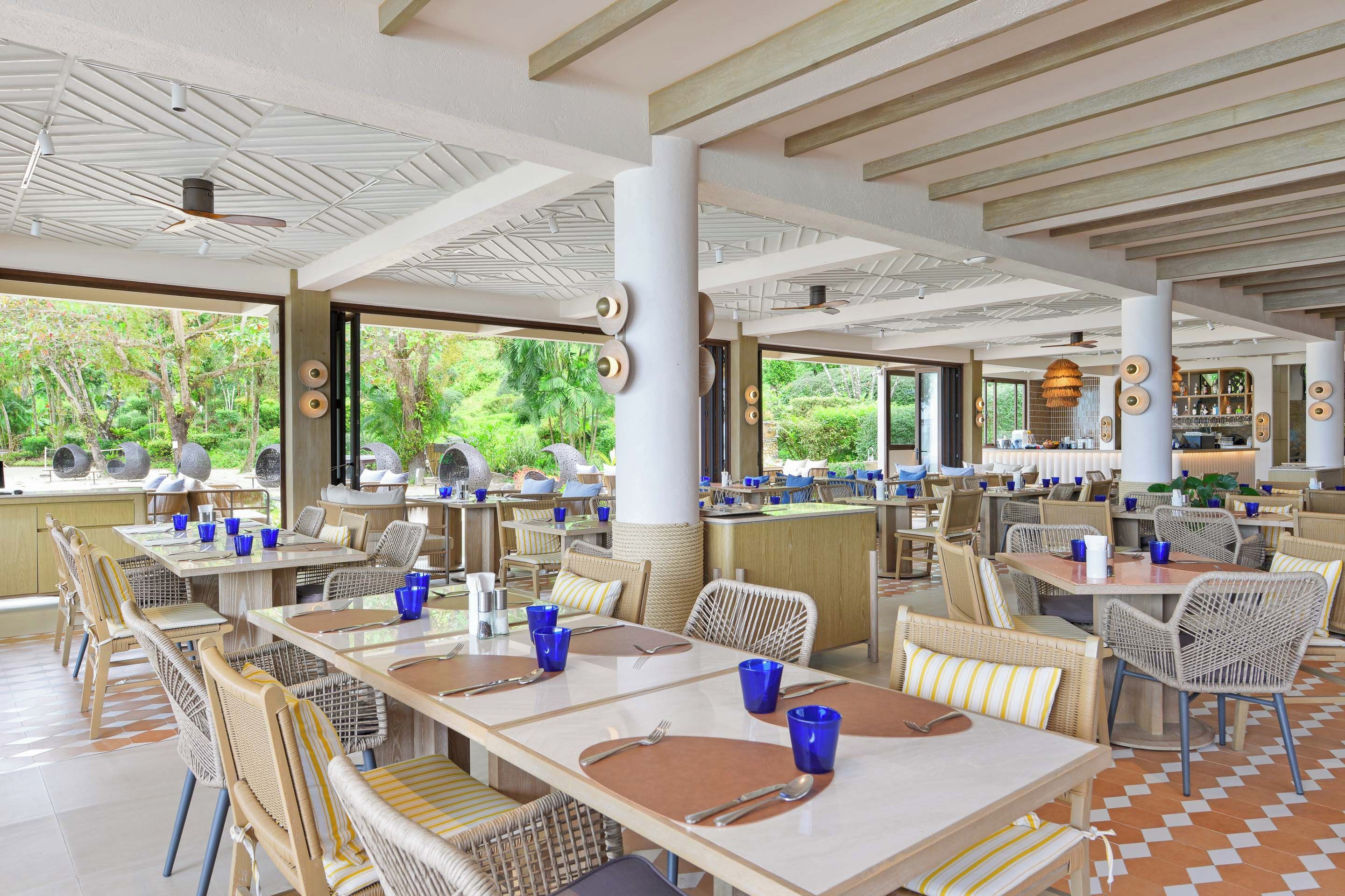 A spacious outdoor dining area with multiple tables and chairs, large windows offering a view of lush greenery, and a ceiling with a geometric pattern.