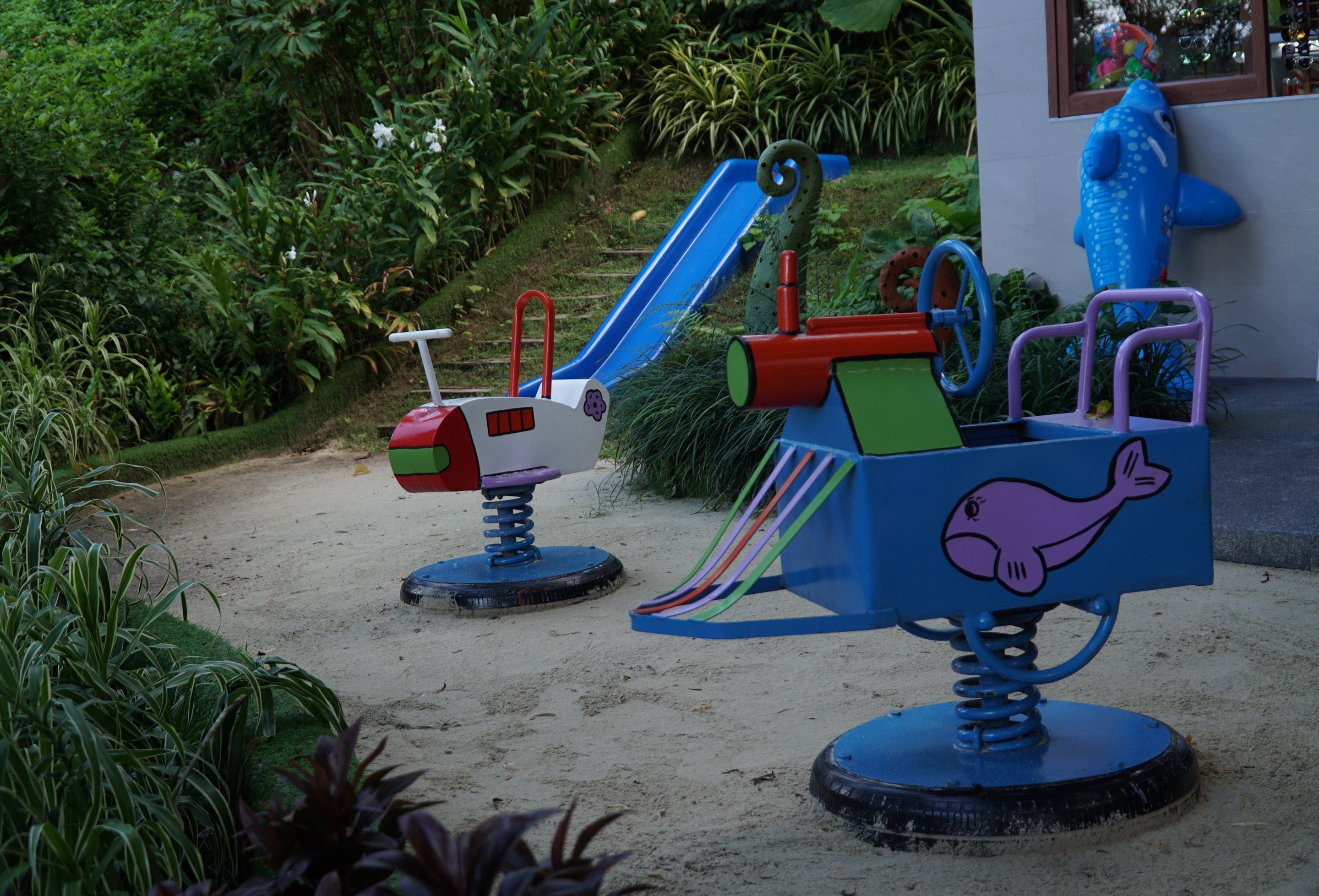 Two children's play equipment with slides and a dolphin design, set on sandy ground with greenery in the background.
