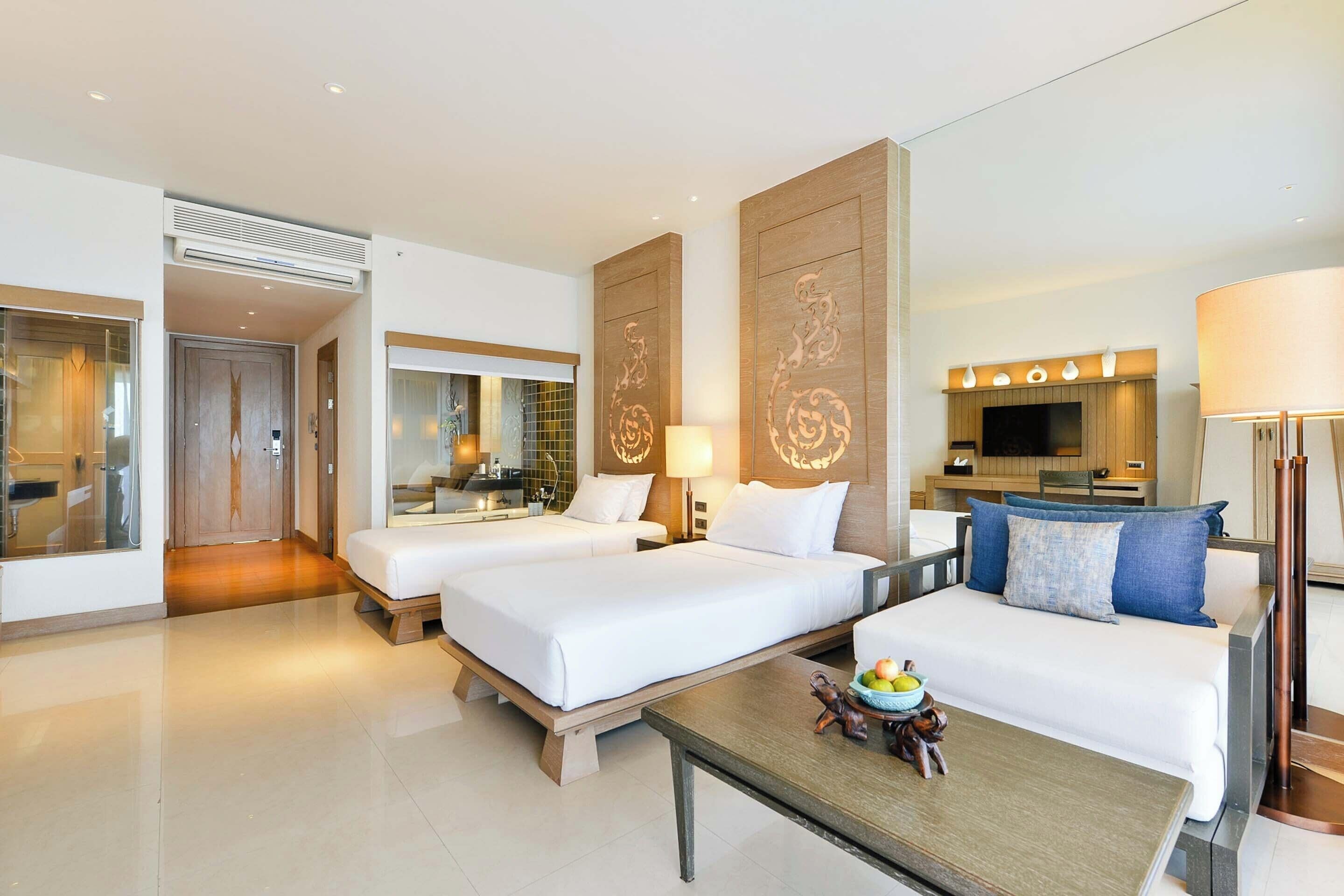 A hotel room with two beds, a sofa, a coffee table, a TV, and a bathroom visible in the background.