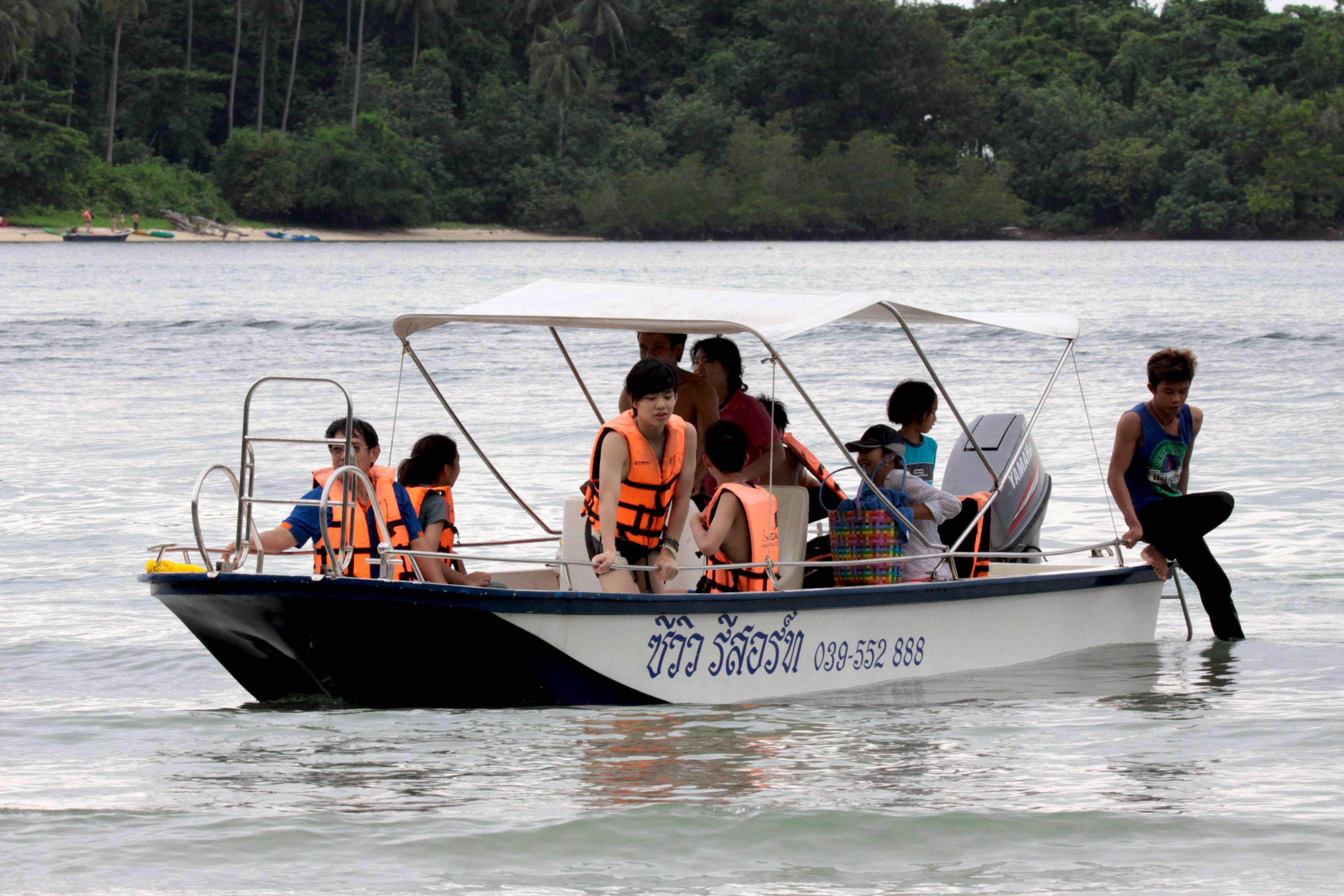 Un bote con pasajeros usando chalecos salvavidas, un timón y un número en el costado.