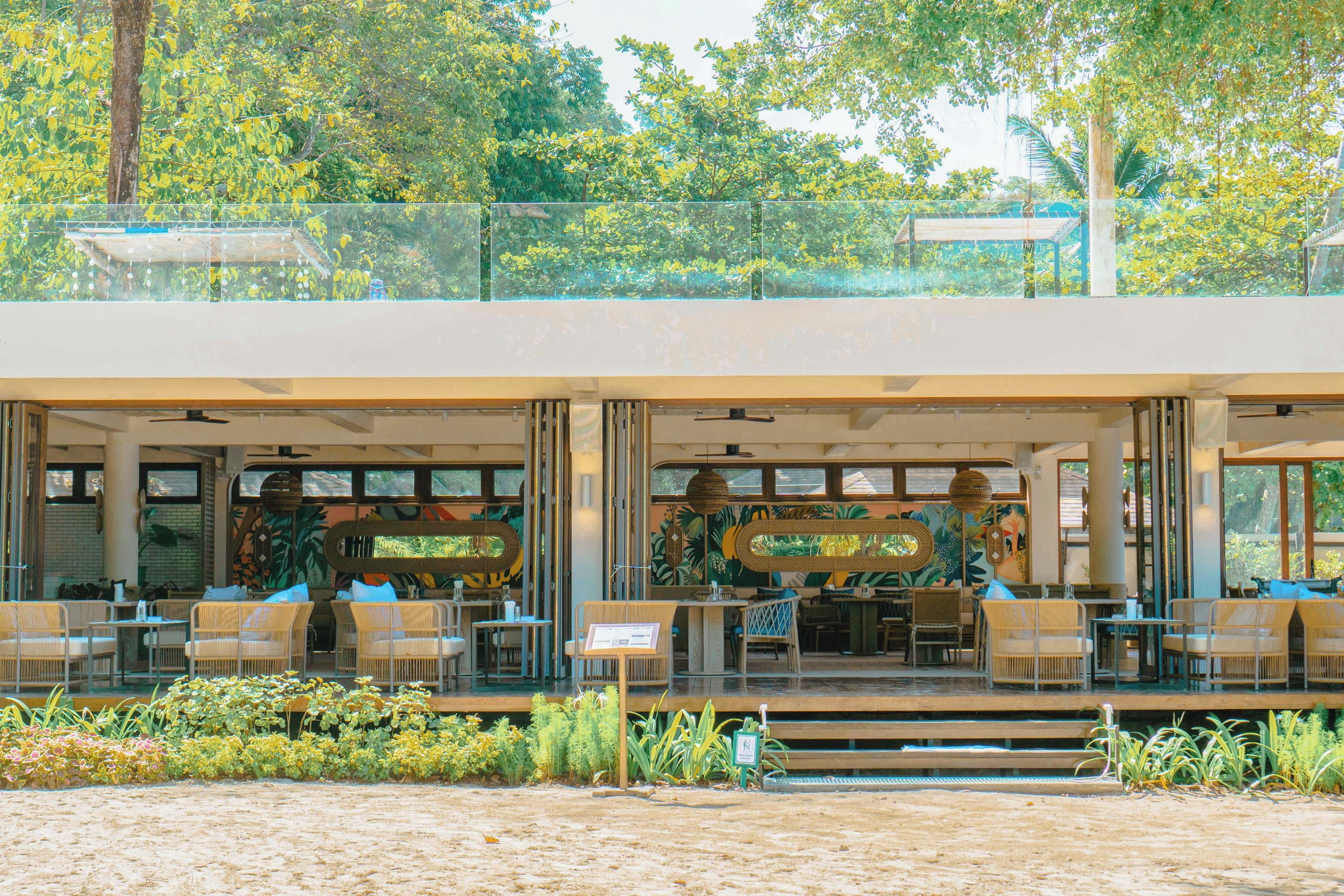 A beachfront restaurant with outdoor seating, a glass balustrade, and tropical murals.