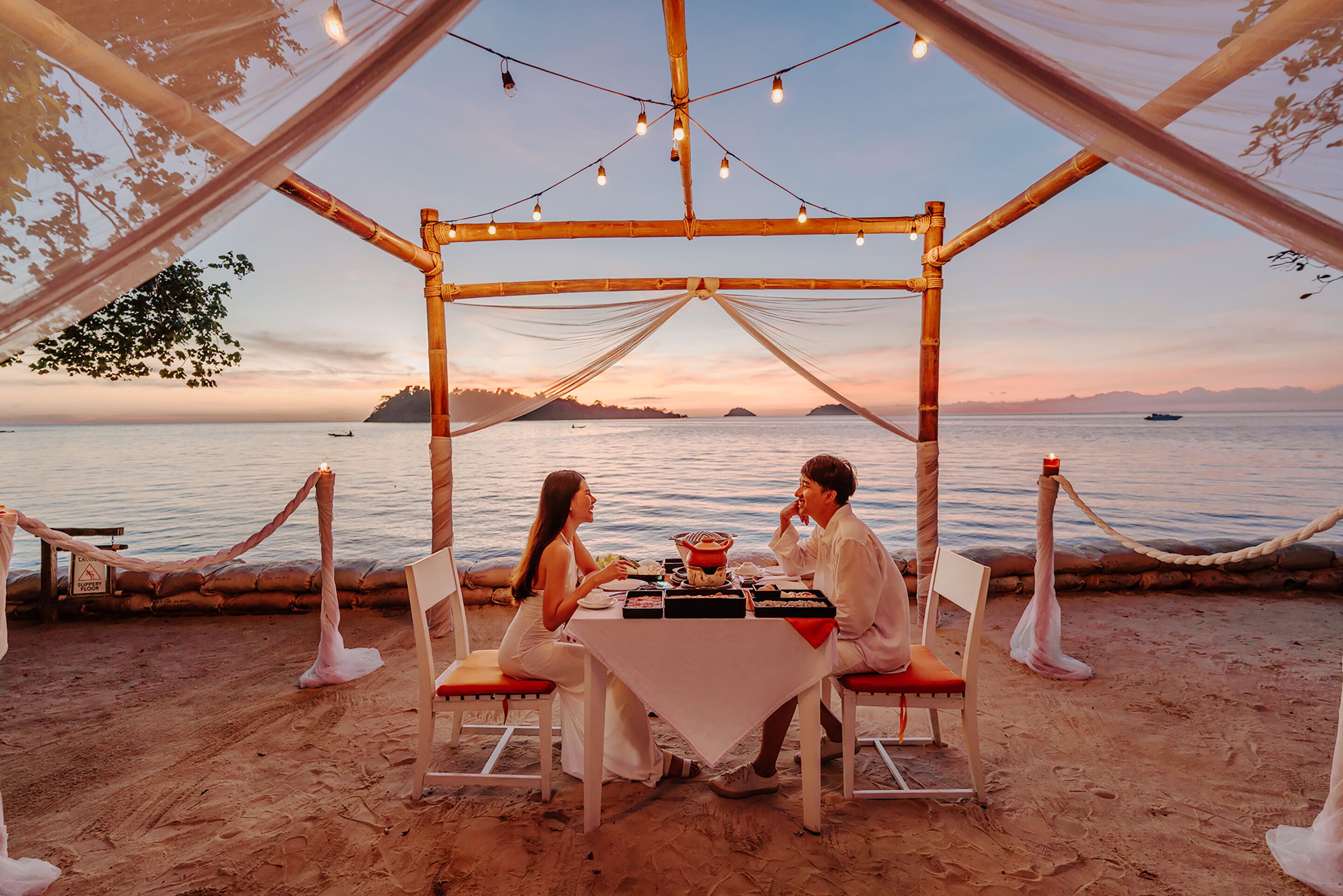 Ein Essbereich am Strand mit Tisch und Stühlen unter einer Bambuskonstruktion mit Blick aufs Meer.