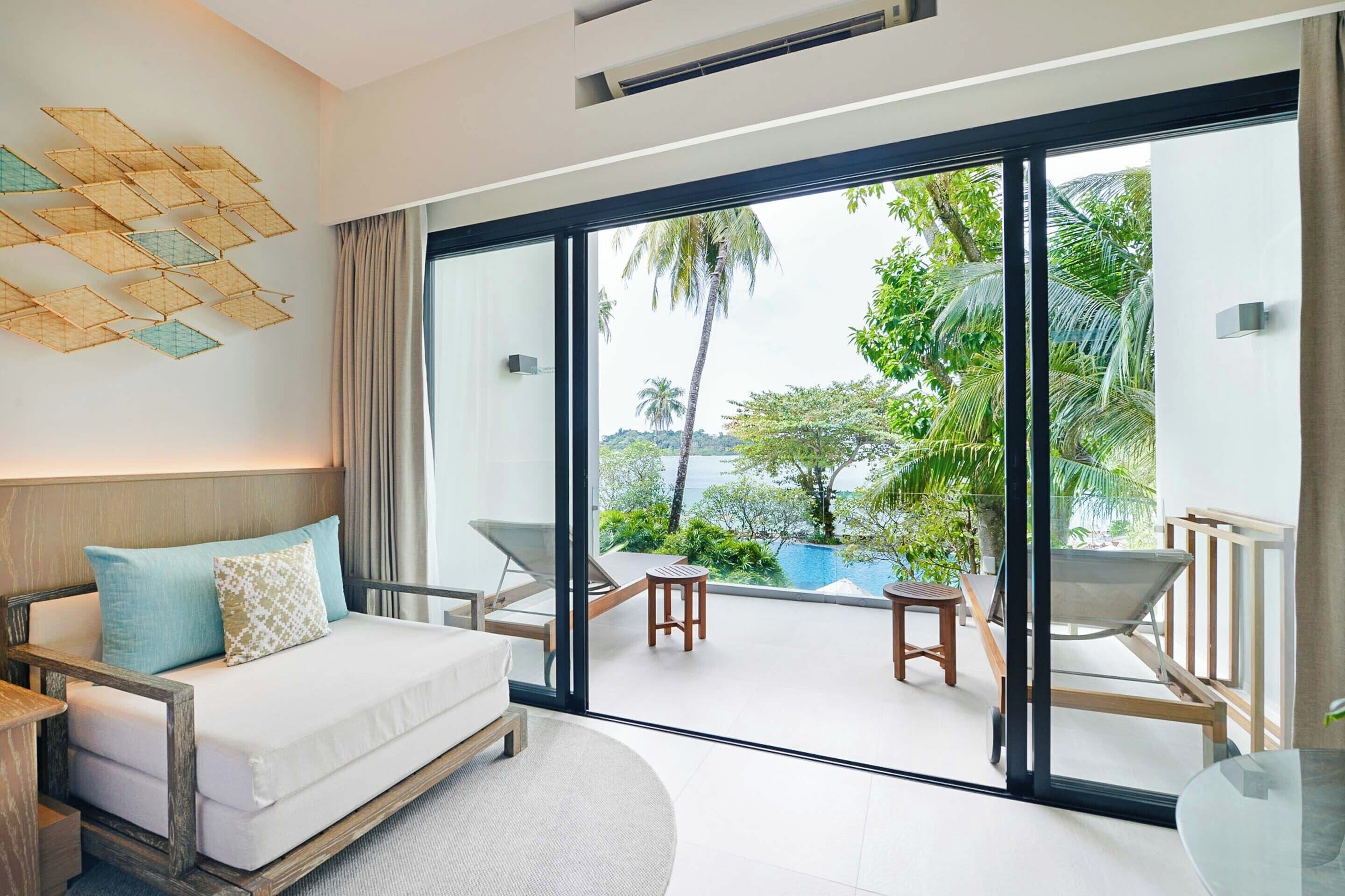 A modern hotel room with a large sliding glass door leading to a balcony with outdoor furniture and a view of greenery.