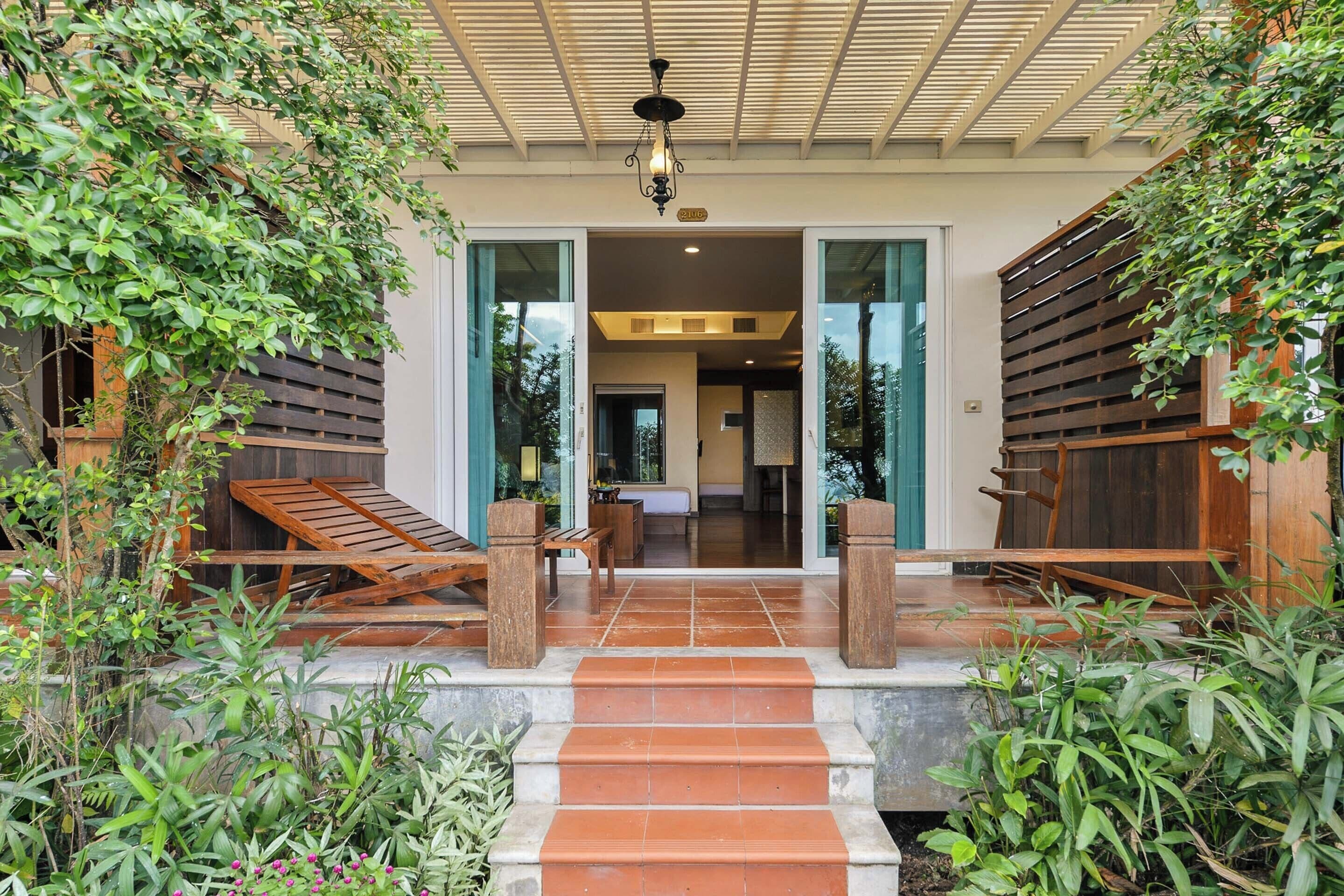 A covered outdoor area with steps leading to a door, surrounded by greenery and wooden furniture.