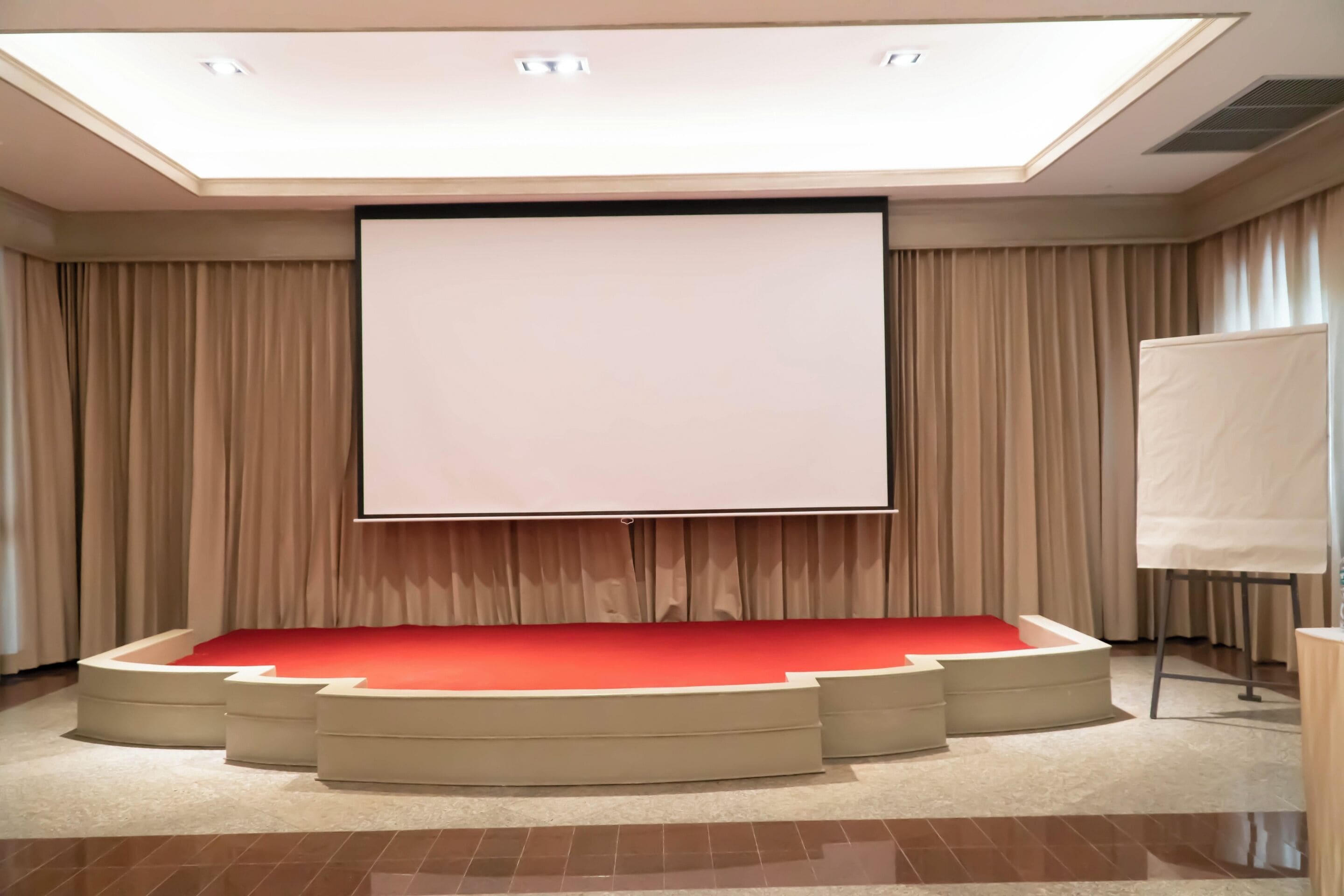 Ein Konferenzraum mit einer großen Leinwand, einer Bühne mit rotem Teppich und Vorhängen.