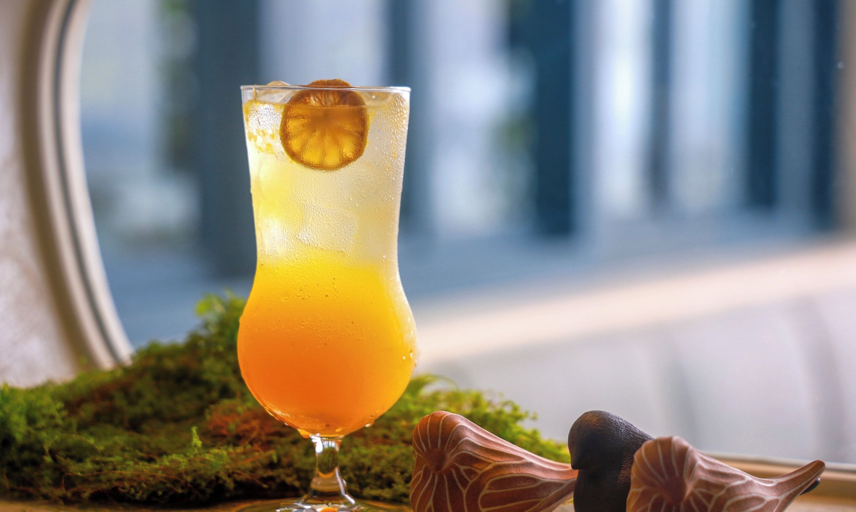 Un grand verre contenant une boisson orange dont la couleur dégrade progressivement, agrémenté d’une tranche d’orange, posé sur une surface recouverte de mousse, avec des objets décoratifs.