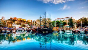 A harbor with multiple boats docked, a building with a red roof, and a stone wall in the background. - Hotel Mevre (Antalya)