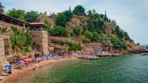 A coastal hotel with a stone facade, a beach with umbrellas, and a rocky cliff. - Hotel Mevre (Antalya)