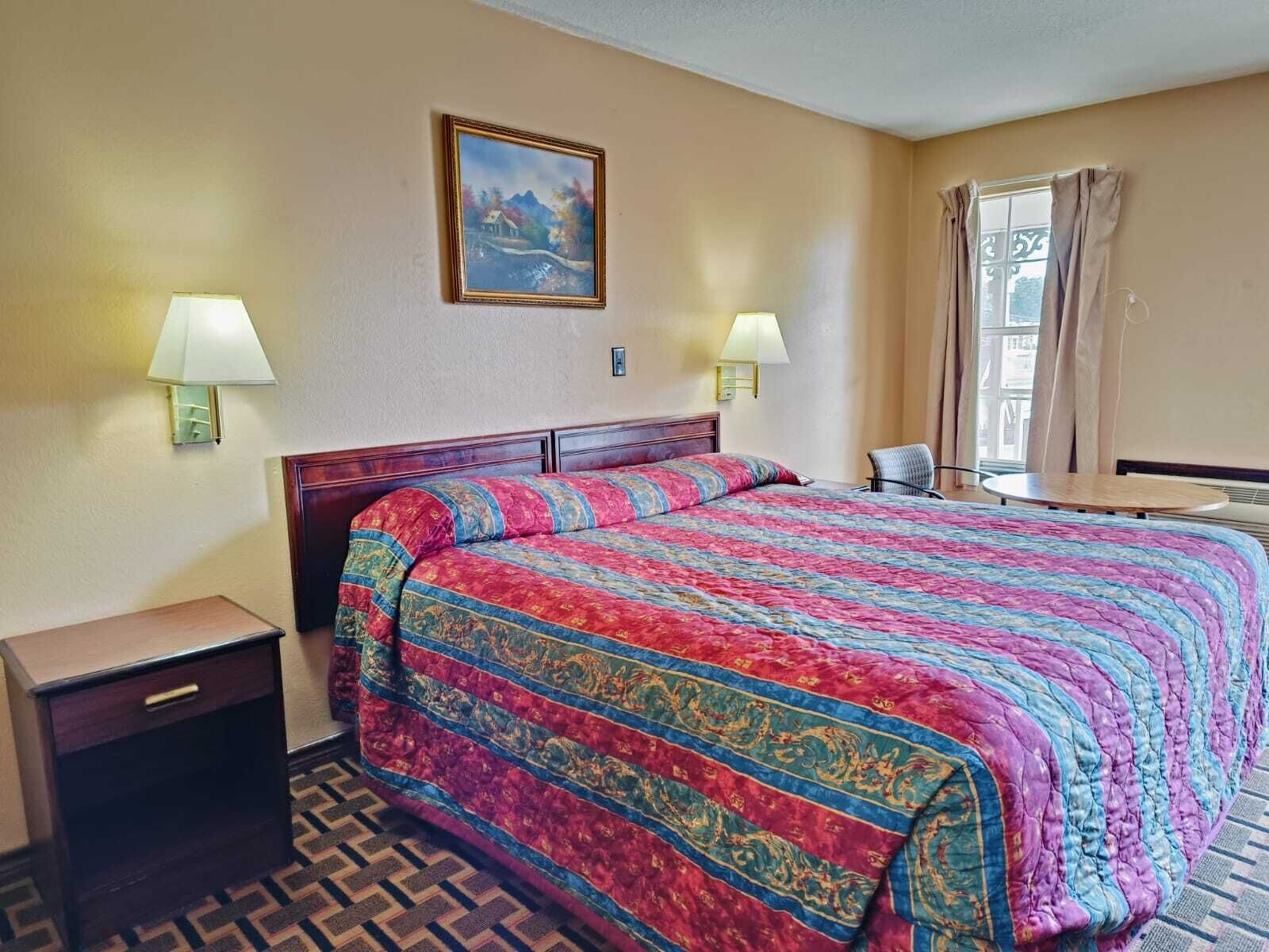 A hotel room with a large bed, two bedside lamps, a nightstand, a painting on the wall, and a window with curtains.