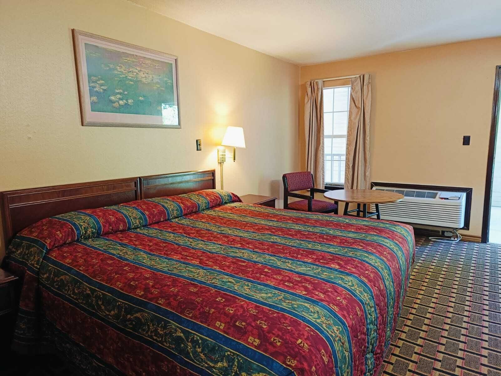 A hotel room with a large bed, a small table, a chair, a window with curtains, and a wall-mounted air conditioner.