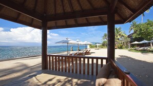 Eine Strandlounge mit Reetdach, gepolsterten Sitzgelegenheiten und Sonnenschirmen.