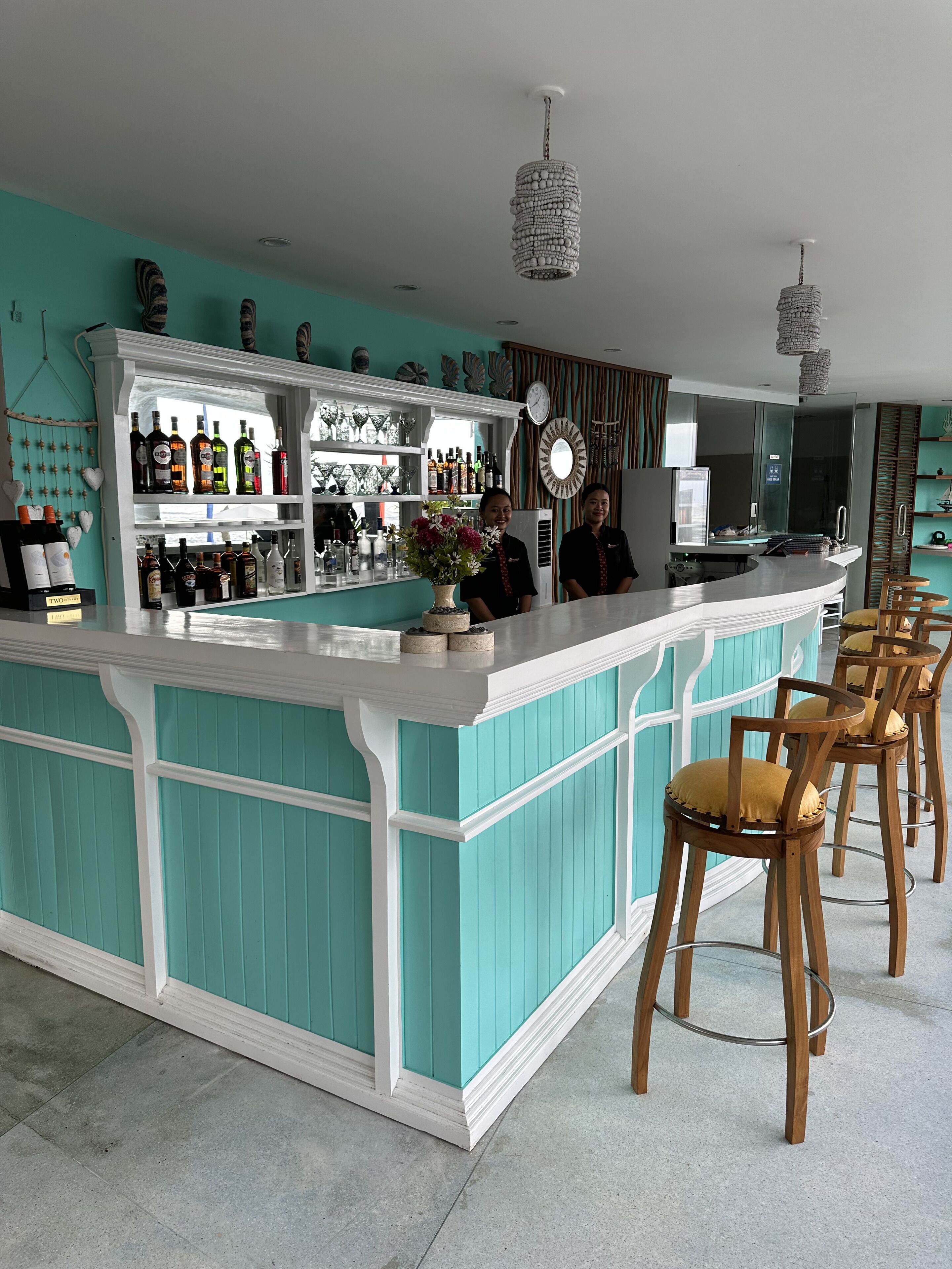 Zona de bar con una decoración color aguamarina, taburetes de madera y una variedad de botellas en estantes.