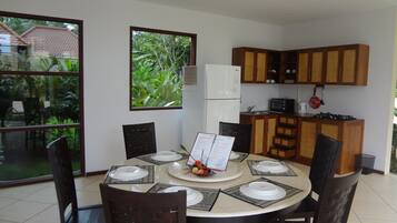 Un coin repas avec une table ronde dressée pour quatre personnes, entourée de chaises. On aperçoit un réfrigérateur et un four à micro-ondes à l’arrière-plan, ainsi que des placards en bois au-dessus du comptoir.
