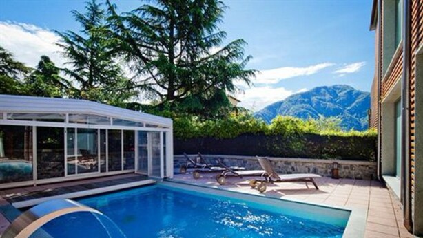 A swimming pool with lounge chairs and a glass-enclosed structure, surrounded by greenery and mountains in the background. - Residence Lenno (Tremezzina)