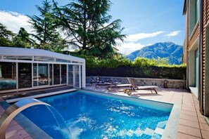 A swimming pool with lounge chairs and a glass-enclosed structure, surrounded by greenery and mountains in the background. - Residence Lenno (Tremezzina)