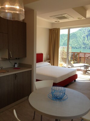A hotel room with a bed, a dining table, a kitchen area, and a balcony with a view of the mountains. - Residence Lenno (Tremezzina)