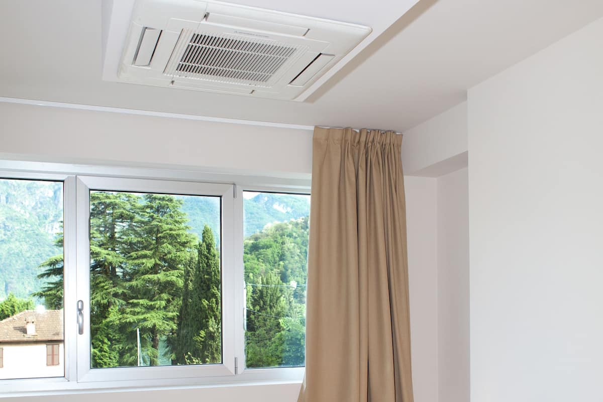 A bedroom with a bed, a window with curtains, a bedside table with a lamp, and a ceiling vent.