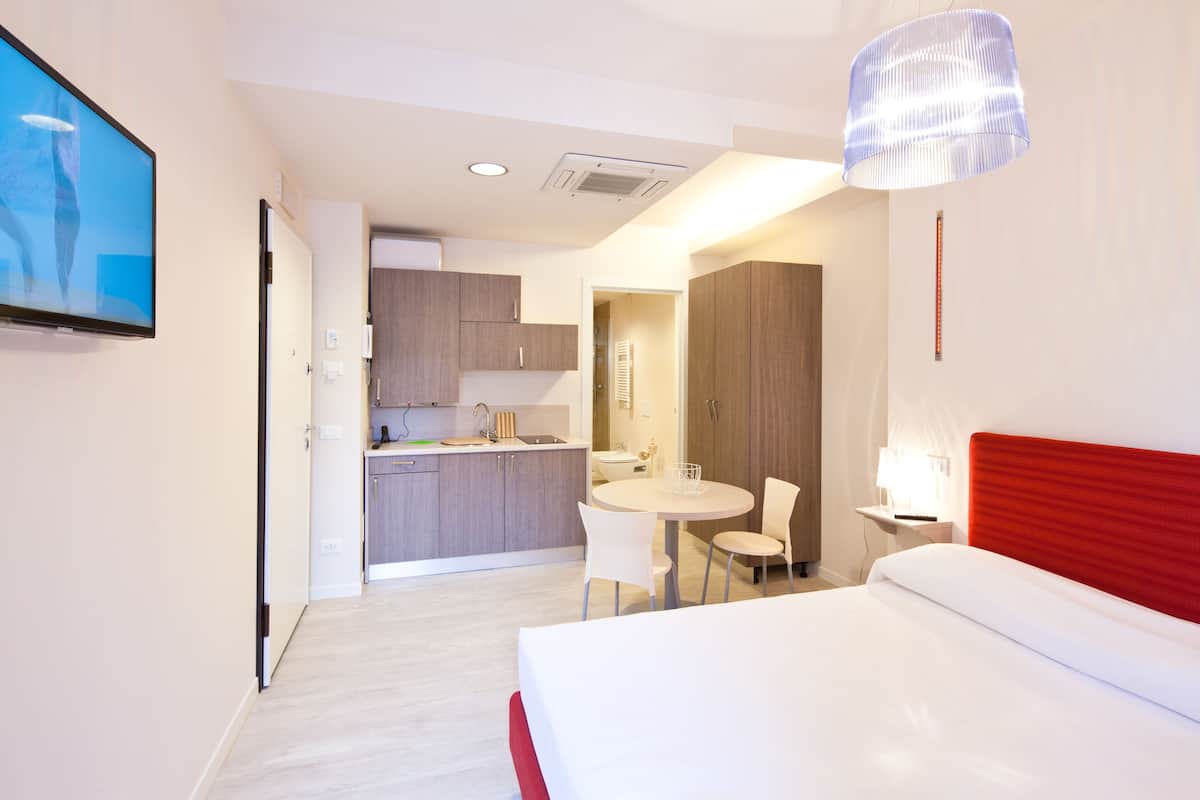 A modern hotel room with a bed, a dining table, and a kitchen area.