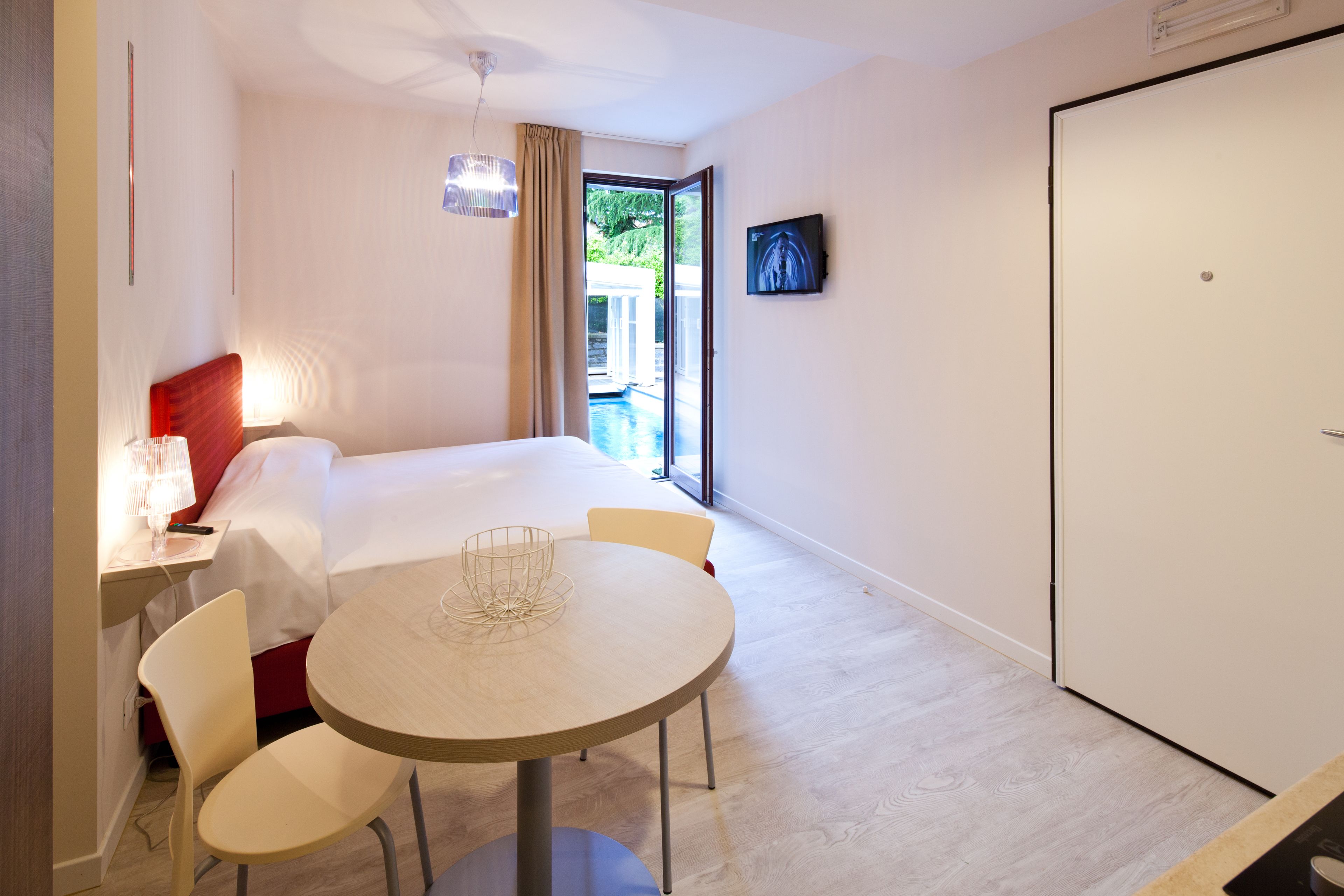 Habitación de hotel con cama, una mesa con sillas, un escritorio con lámpara, televisor en la pared y puerta corrediza al exterior.